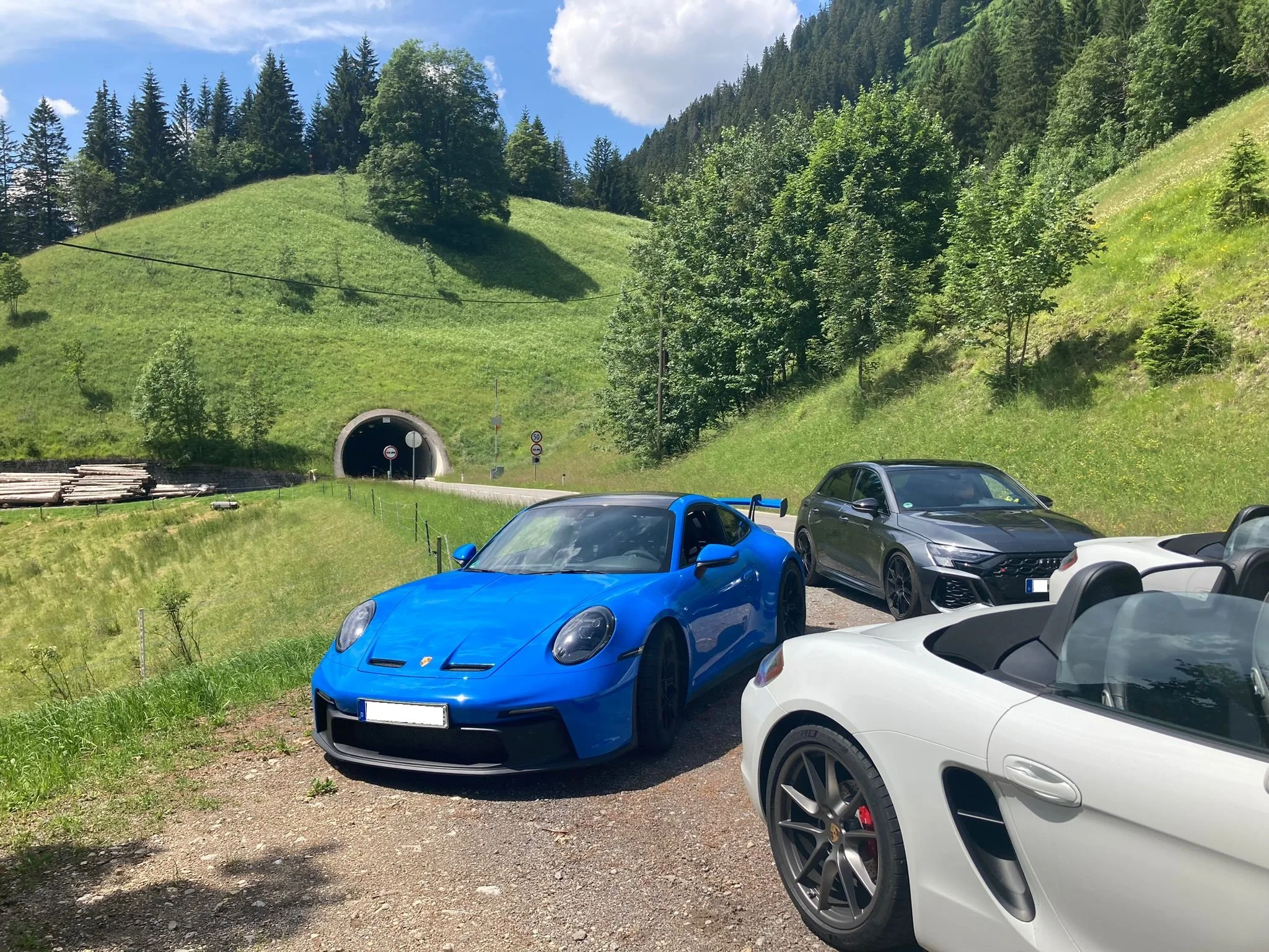 Drei sportliche Autos, darunter ein blauer Porsche, auf einem Parkplatz vor einem Tunnel in einer bergigen Landschaft mit grünen Hängen und Bäumen, bei sonnigem Wetter.