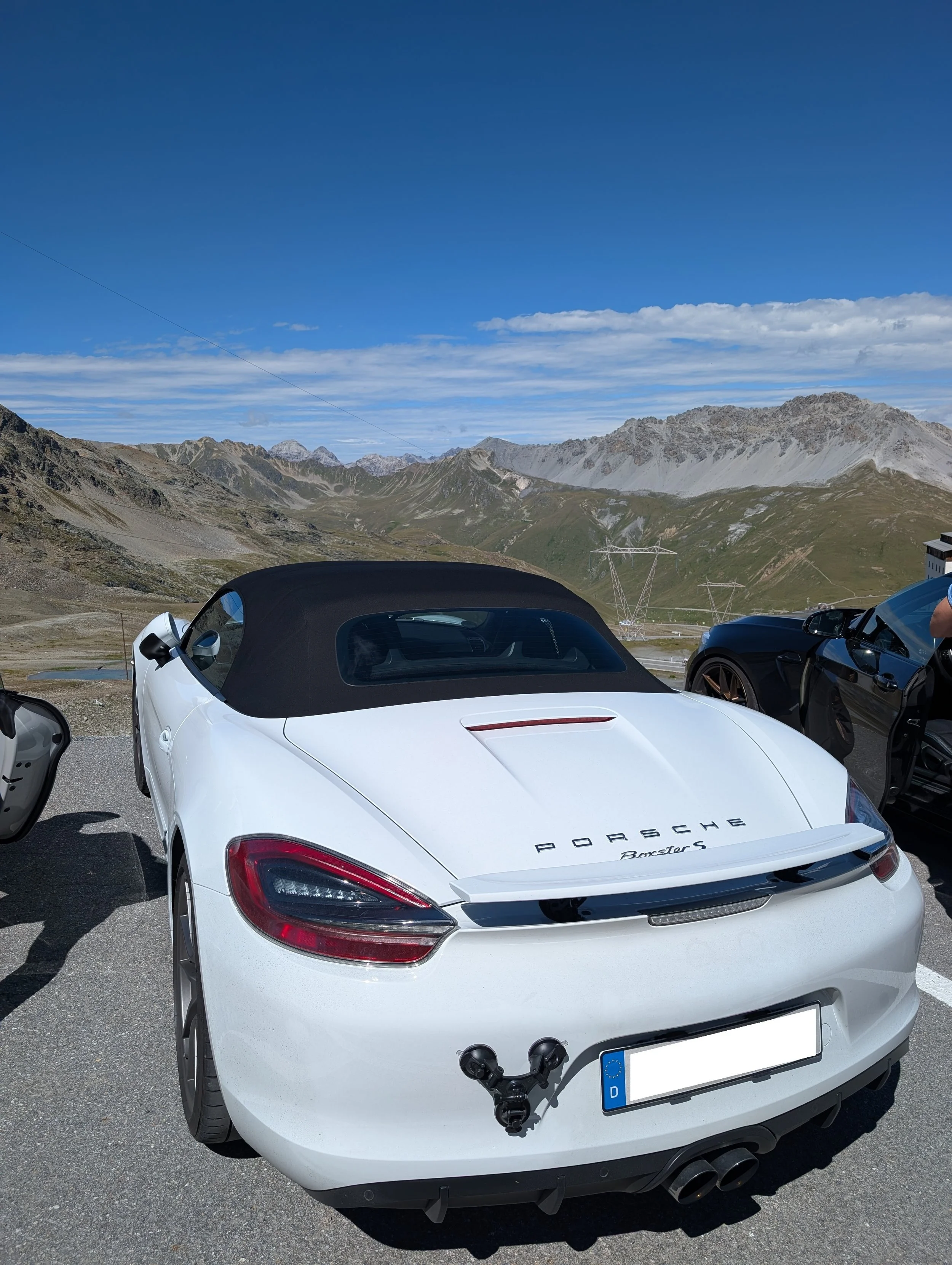Weißer Porsche Boxster S mit schwarzem Verdeck in den Alpen, umgeben von Bergen bei schönem Wetter.
