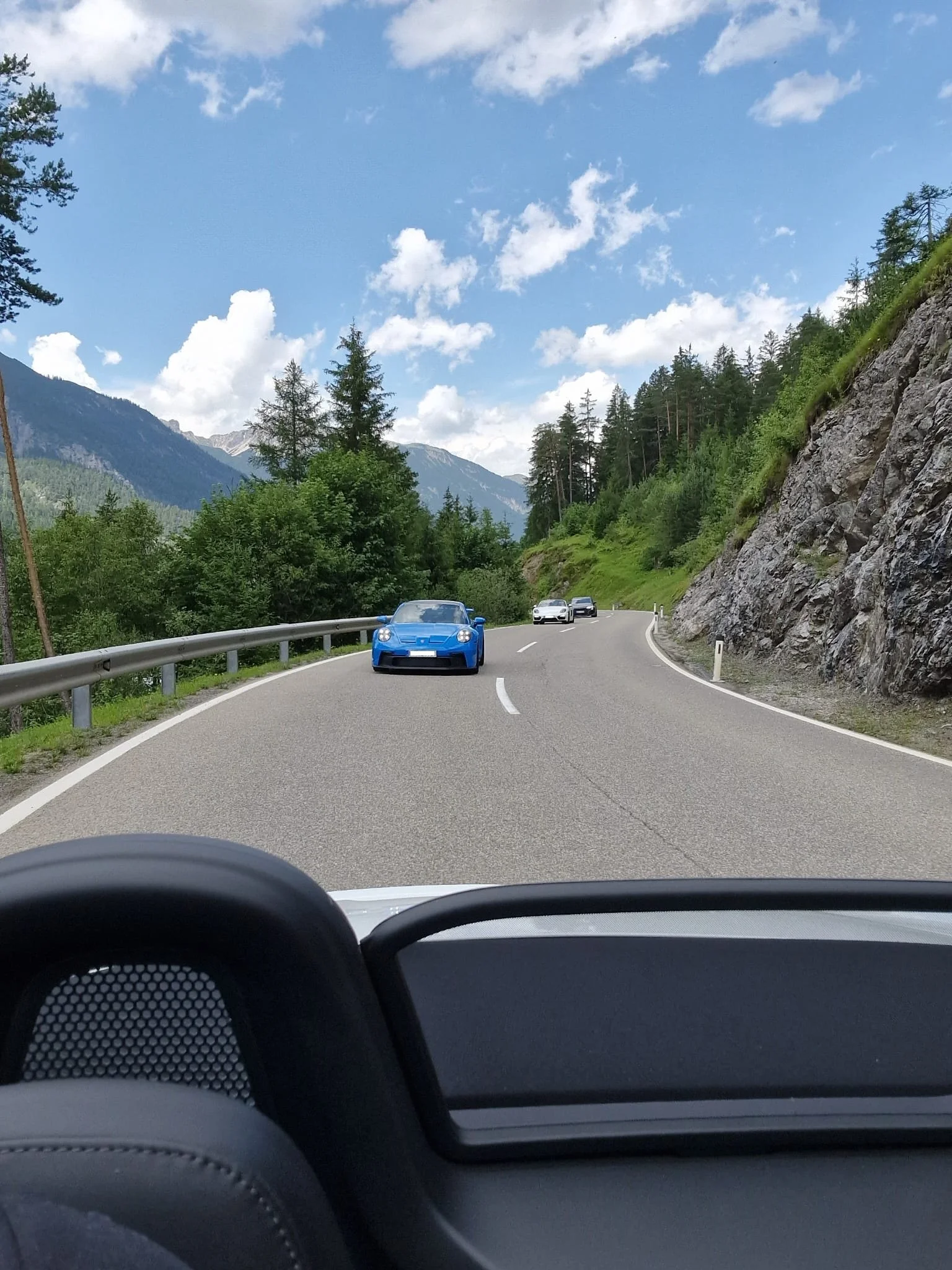 Blick von einem Fahrzeug auf eine kurvige, bergige Straße mit mehreren Autos, umgeben von grünen Bäumen und Felsen, bei gutem Wetter mit blauen Himmel und Wolken.