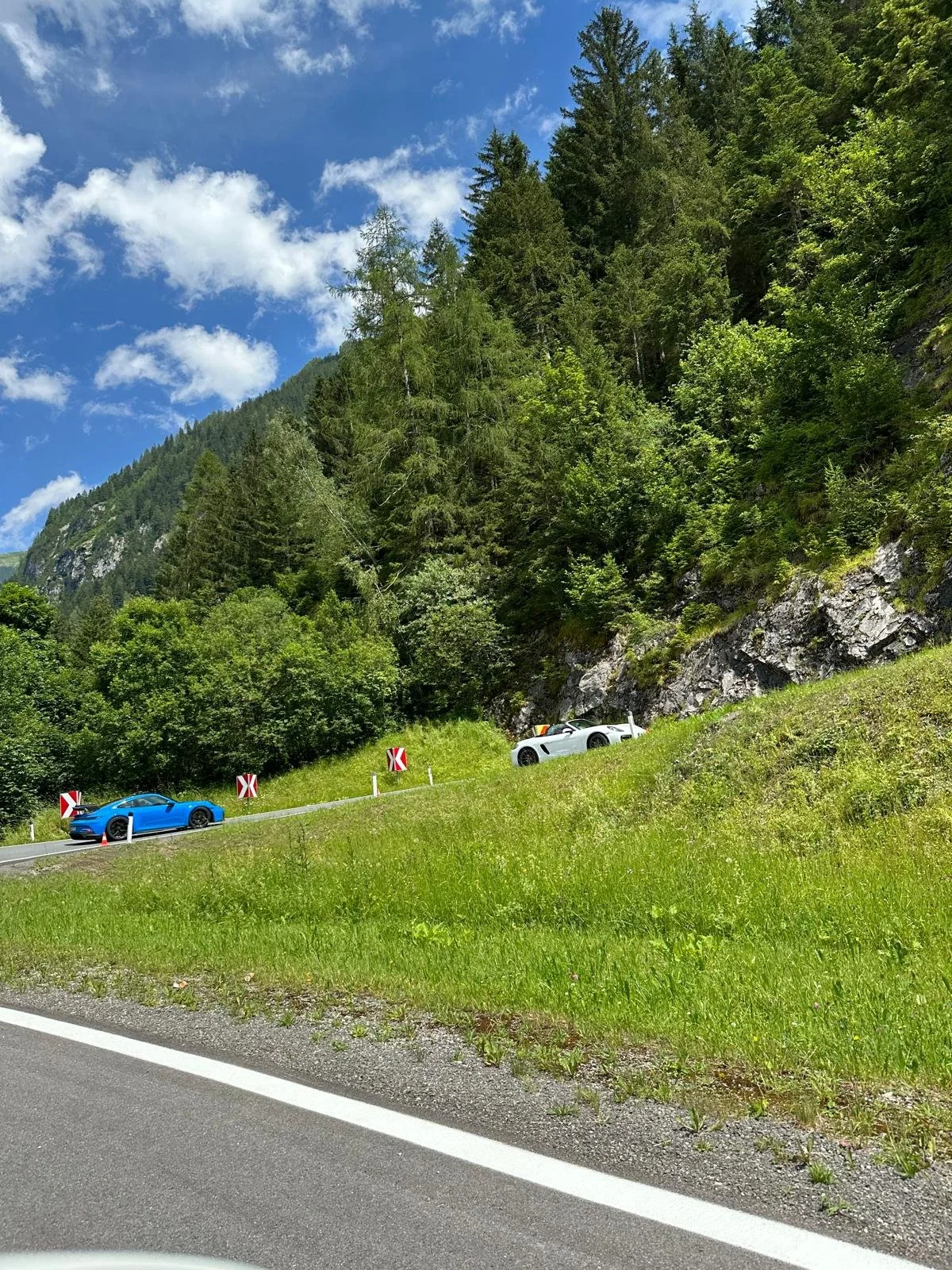 Berglandschaft mit grünen Bäumen, einem blauen Himmel und zwei Sportwagen auf der Straße.