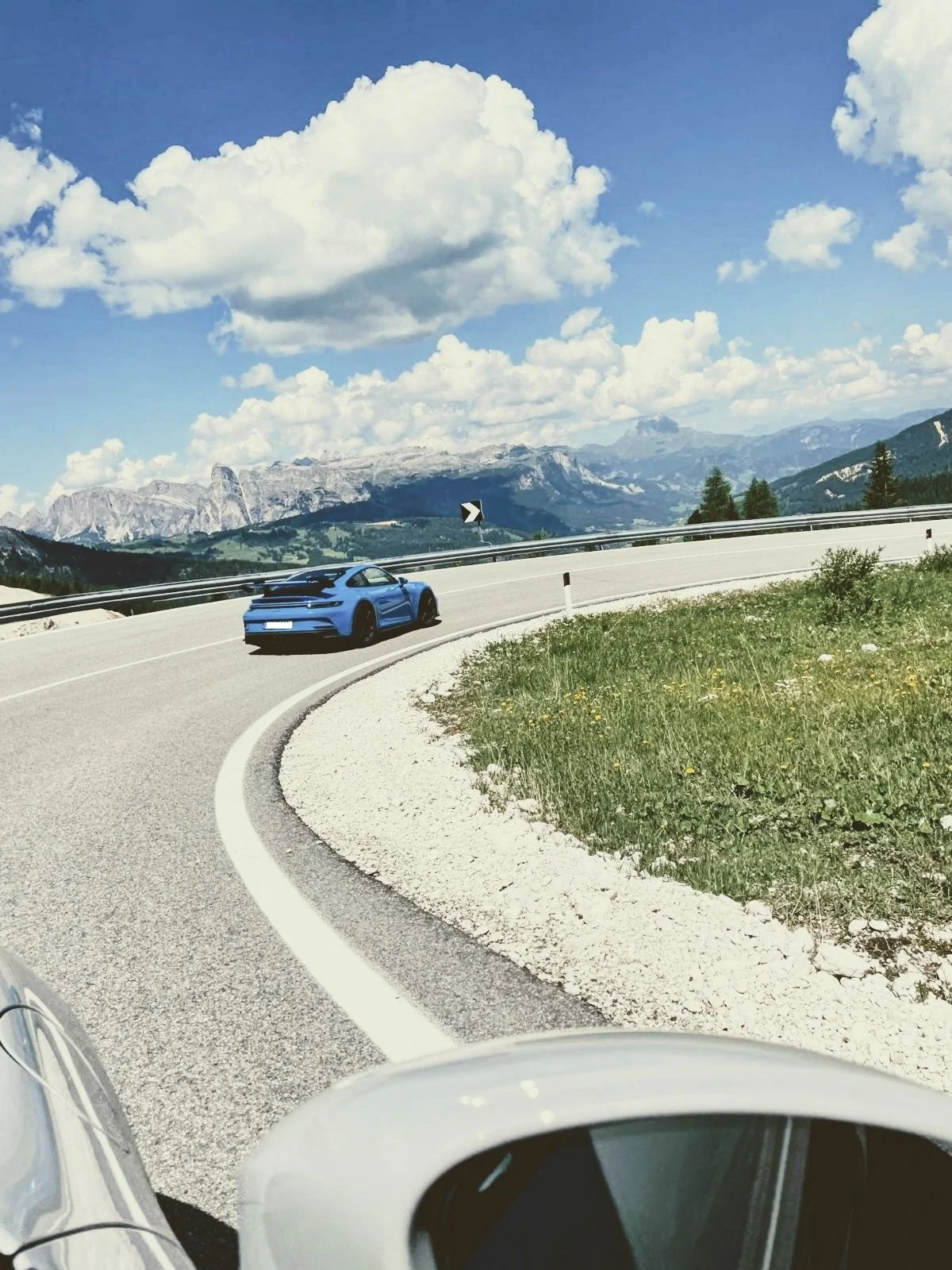 Bergige Landschaft mit kurvigem Straßenabschnitt, blauer Sportwagen und bewölktem Himmel.