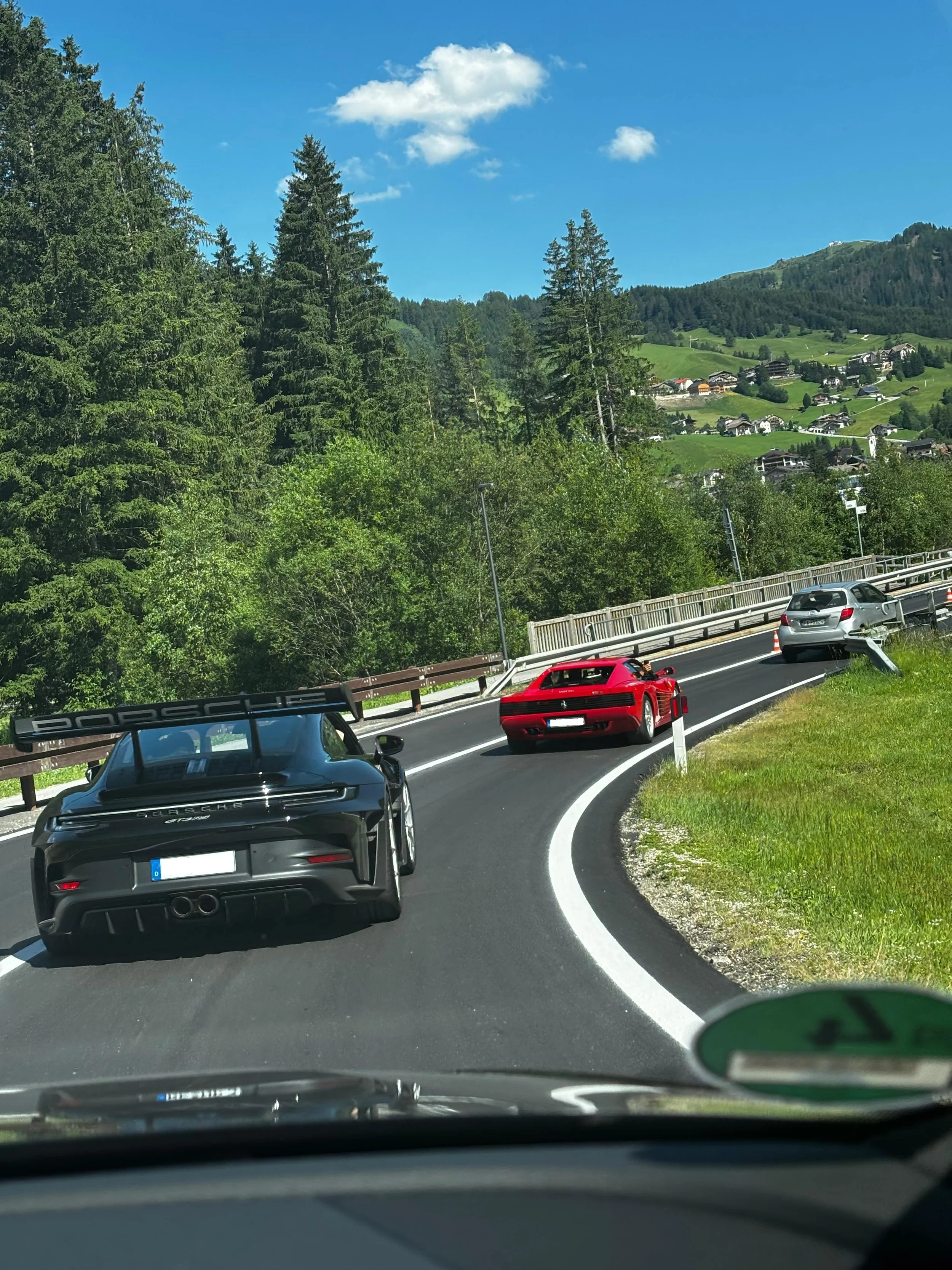 Schnelle Porsche-Sportwagen, die eine kurvige Bergstraße in einer ländlichen Gegend mit grünen Hügeln und Häusern im Hintergrund befahren, während das Bild aus einem Auto heraus aufgenommen wurde.