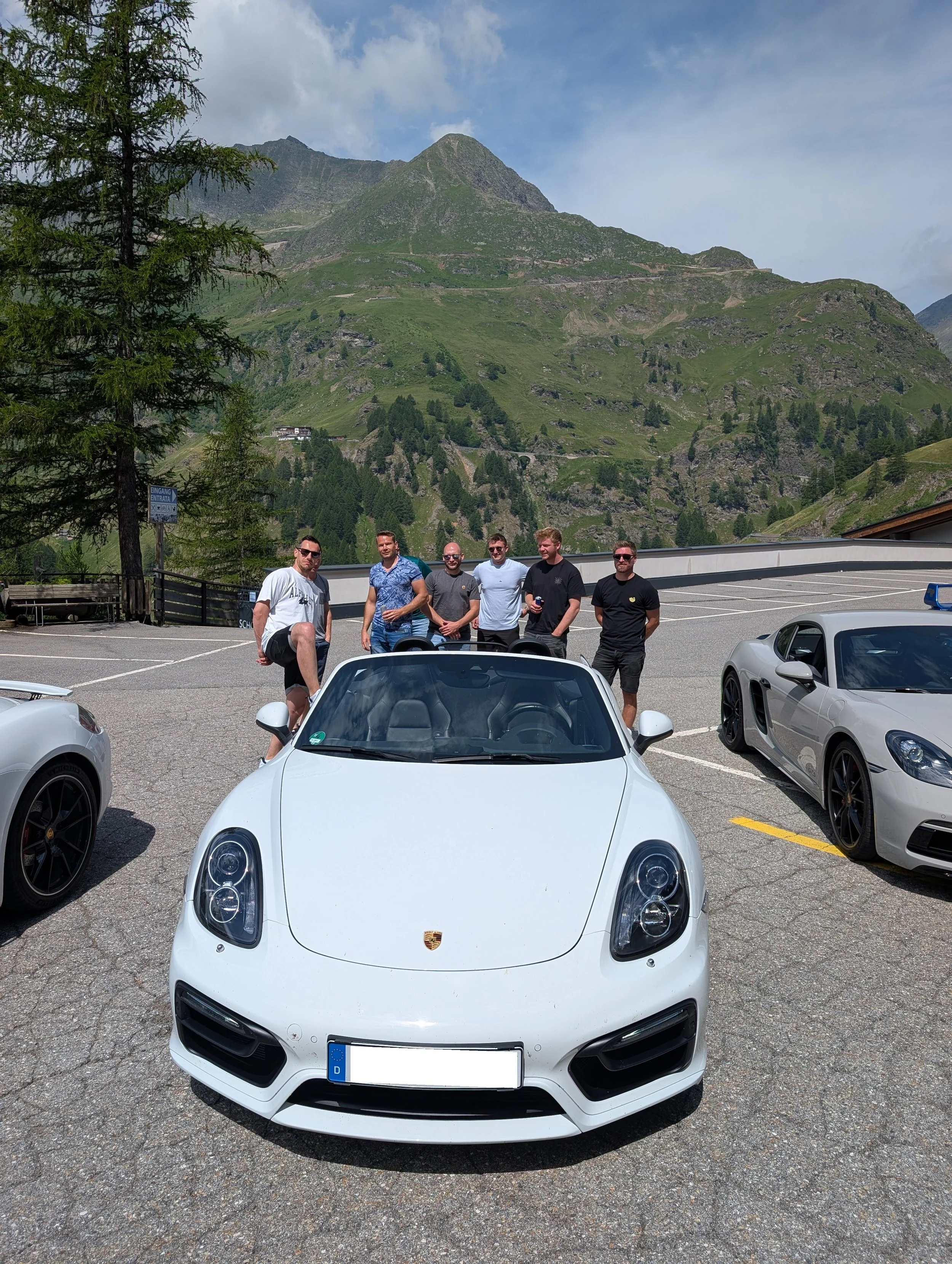 Sechs Männer stehen hinter einem weißen Porsche Roadster in einer Parklücke mit Blick auf grüne Berge und bewölkten Himmel, umgeben von weiteren Sportwagen in einer Bergregion.