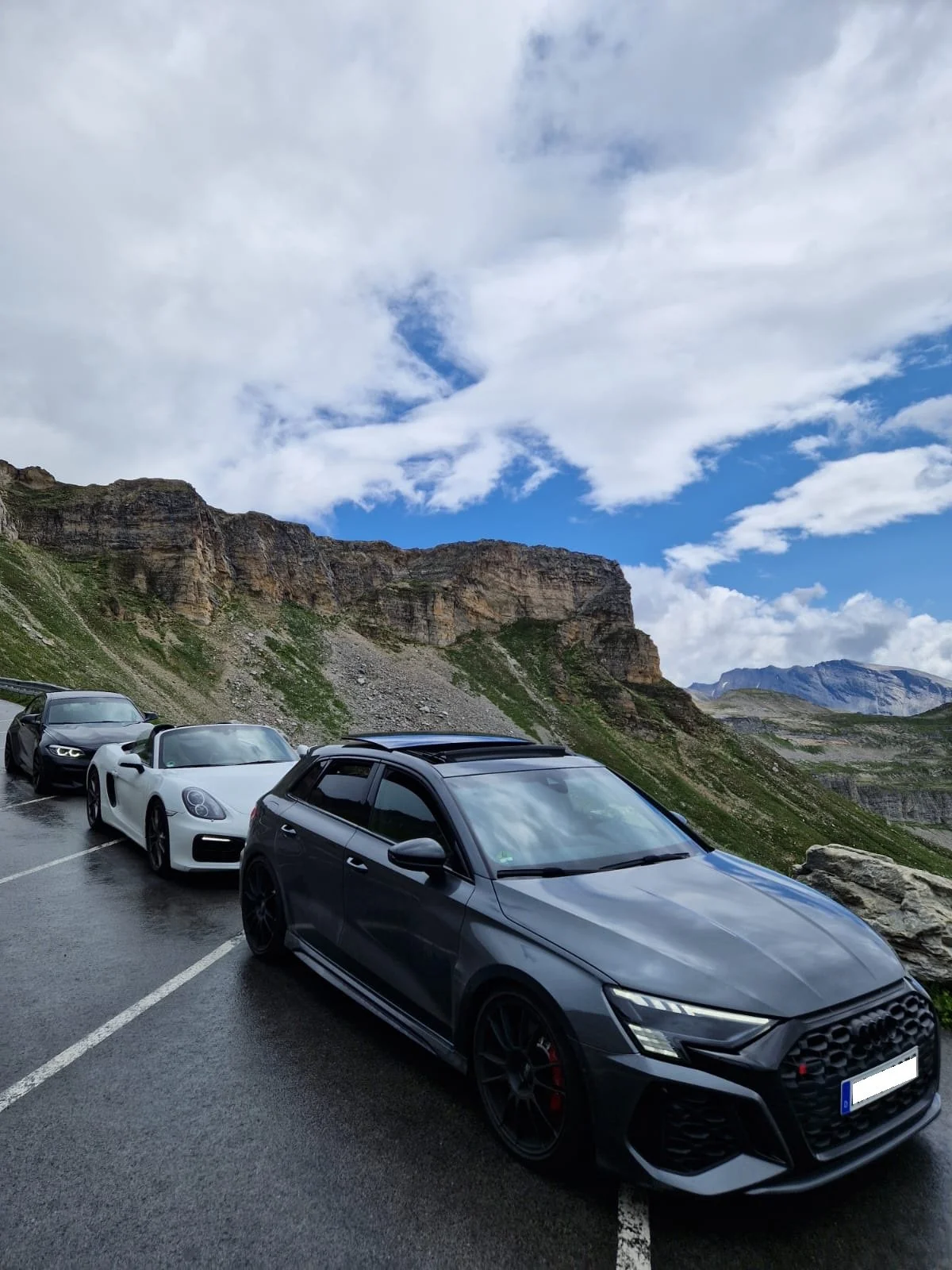 Vier Autos parken an einer Bergstraße mit steilem, felsigem Hintergrund und bewölktem Himmel.