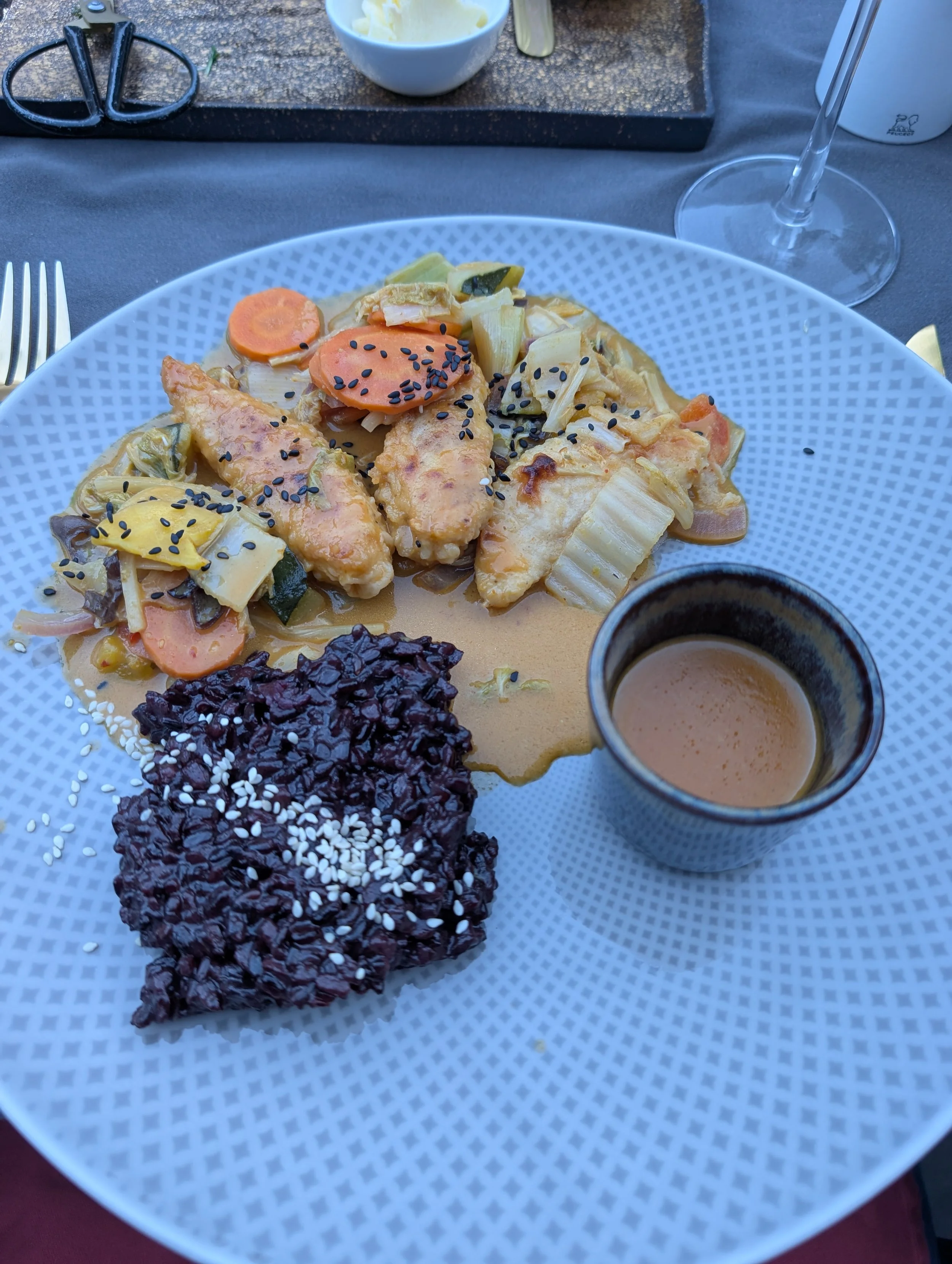 Bunte Mahlzeit mit Hähnchen, Karotten, Zucchini, Reis, schwarze Bohnen mit Sesam und Sauce auf einem weißen Teller, Getränke, Besteck und weitere Speisen im Hintergrund.