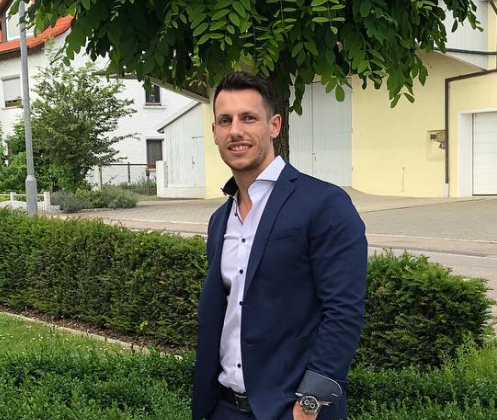 Ein Mann in einem dunklen Anzug steht im Freien vor Grünflächen und Häusern, lächelt in die Kamera.