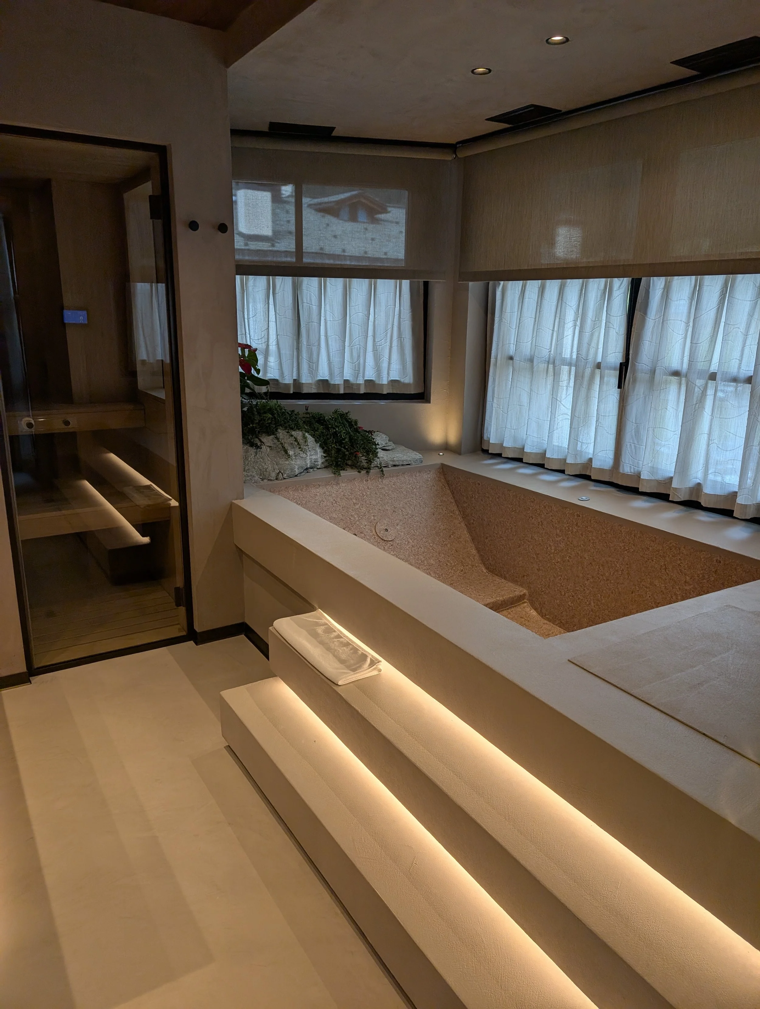 Ein Badezimmer mit einer Badewanne, weißen Vorhängen vor den Fenstern, und einem Saunaraum mit Glastür, Holzbank und Beleuchtung.