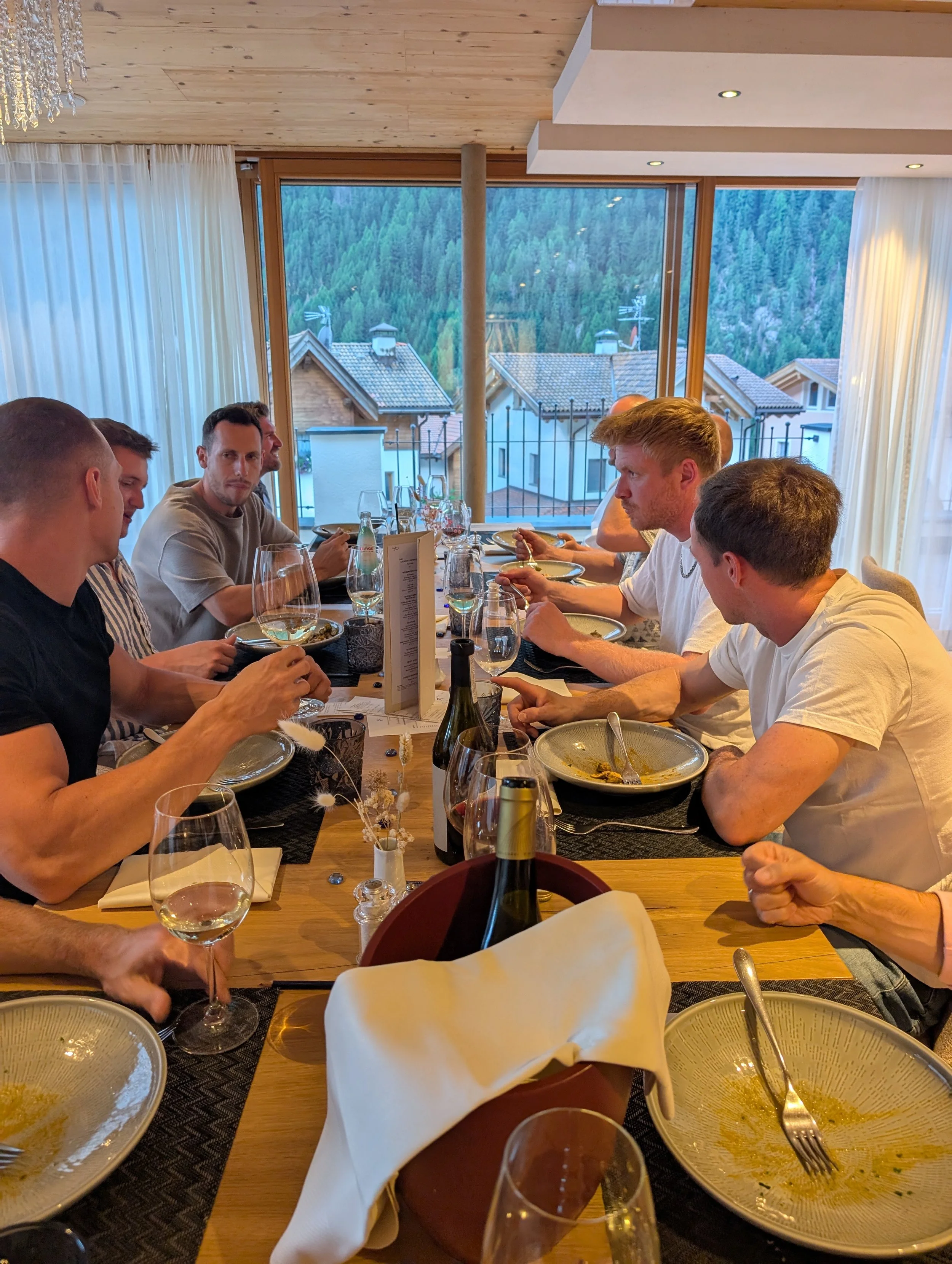 Menschen sitzen an einem Esstisch in einem Restaurant, essen und unterhalten sich, mit großen Fenstern im Hintergrund, die einen Berg mit Bäumen zeigen.