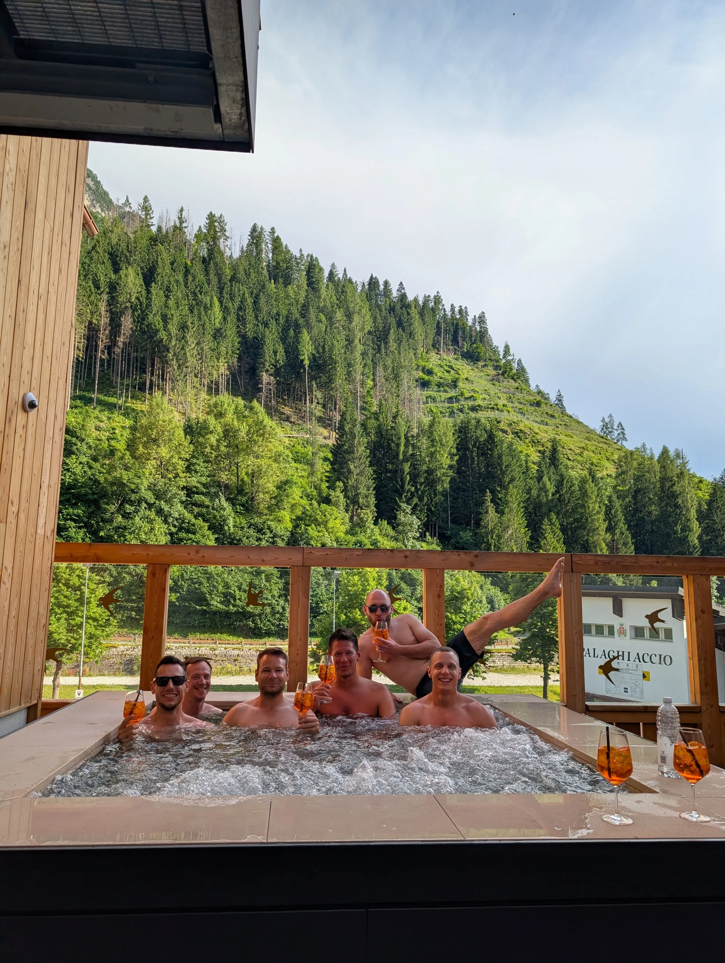 Fünf Männer und ein Mann in Aquafitness-Posen sitzen in einem Whirlpool im Freien, trinken Getränke, mit einer Berg- und Waldkulisse im Hintergrund, bei sonnigem Wetter.