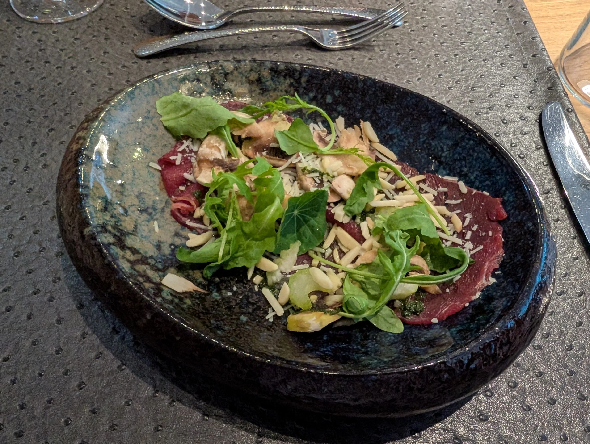 Rindersalat mit Rucola, gehobeltem Käse, Pilzen und Nüssen serviert auf einer schwarzen Steinschale.