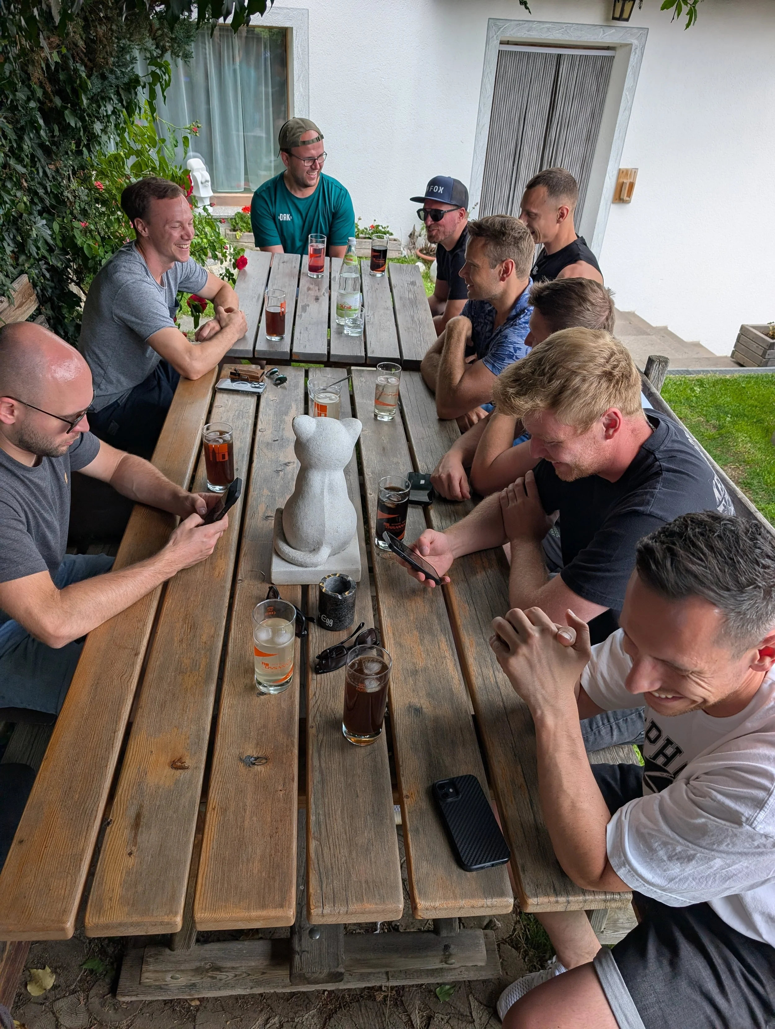 Eine Gruppe junger Männer sitzt an einem langen Holztisch im Freien, unterhält sich und schaut auf ihre Handys, Getränke vor ihnen.