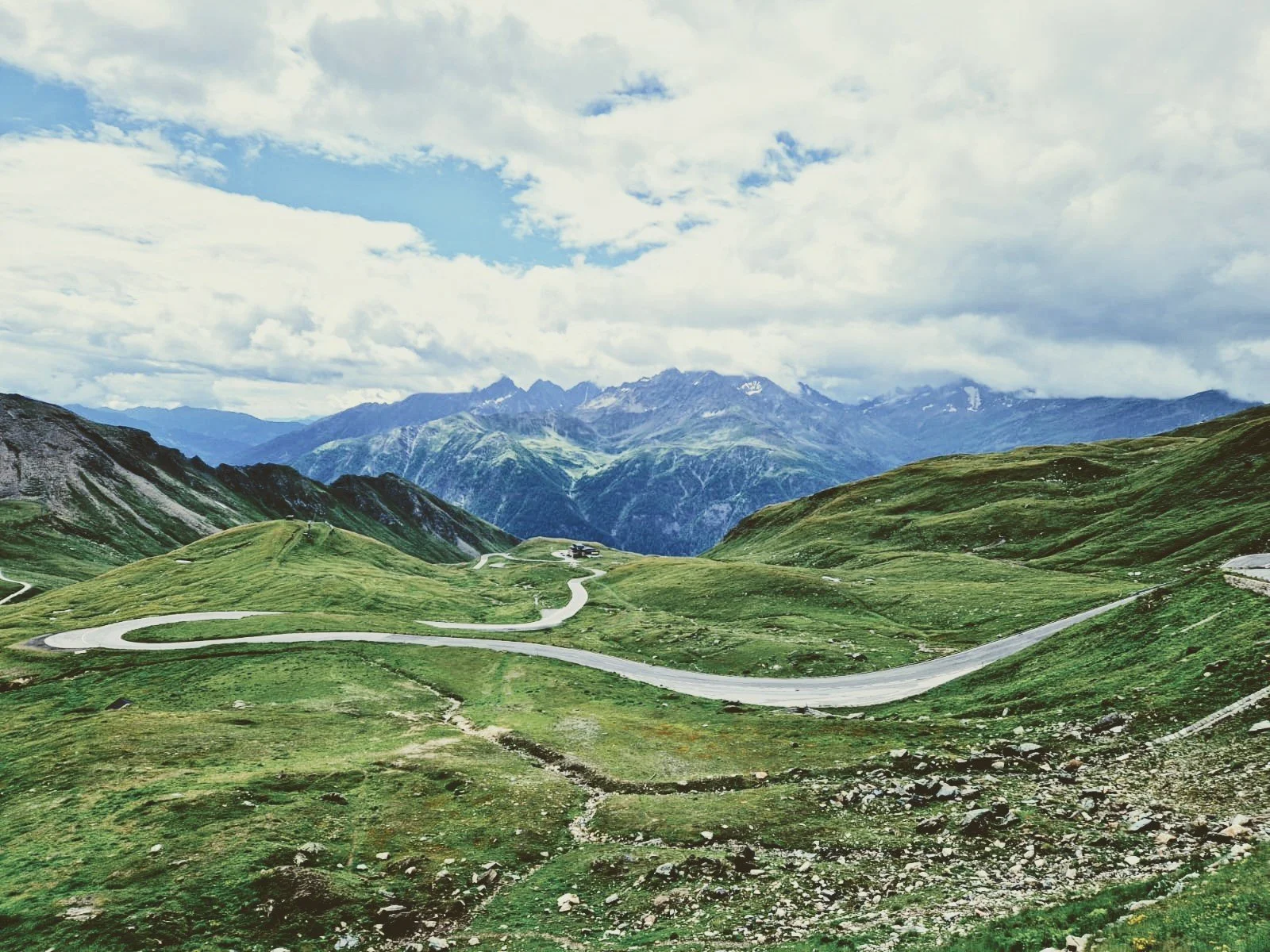 Eine grüne Hügellandschaft mit kurviger Straße, im Hintergrund Berge und eine bewölkte Himmel.