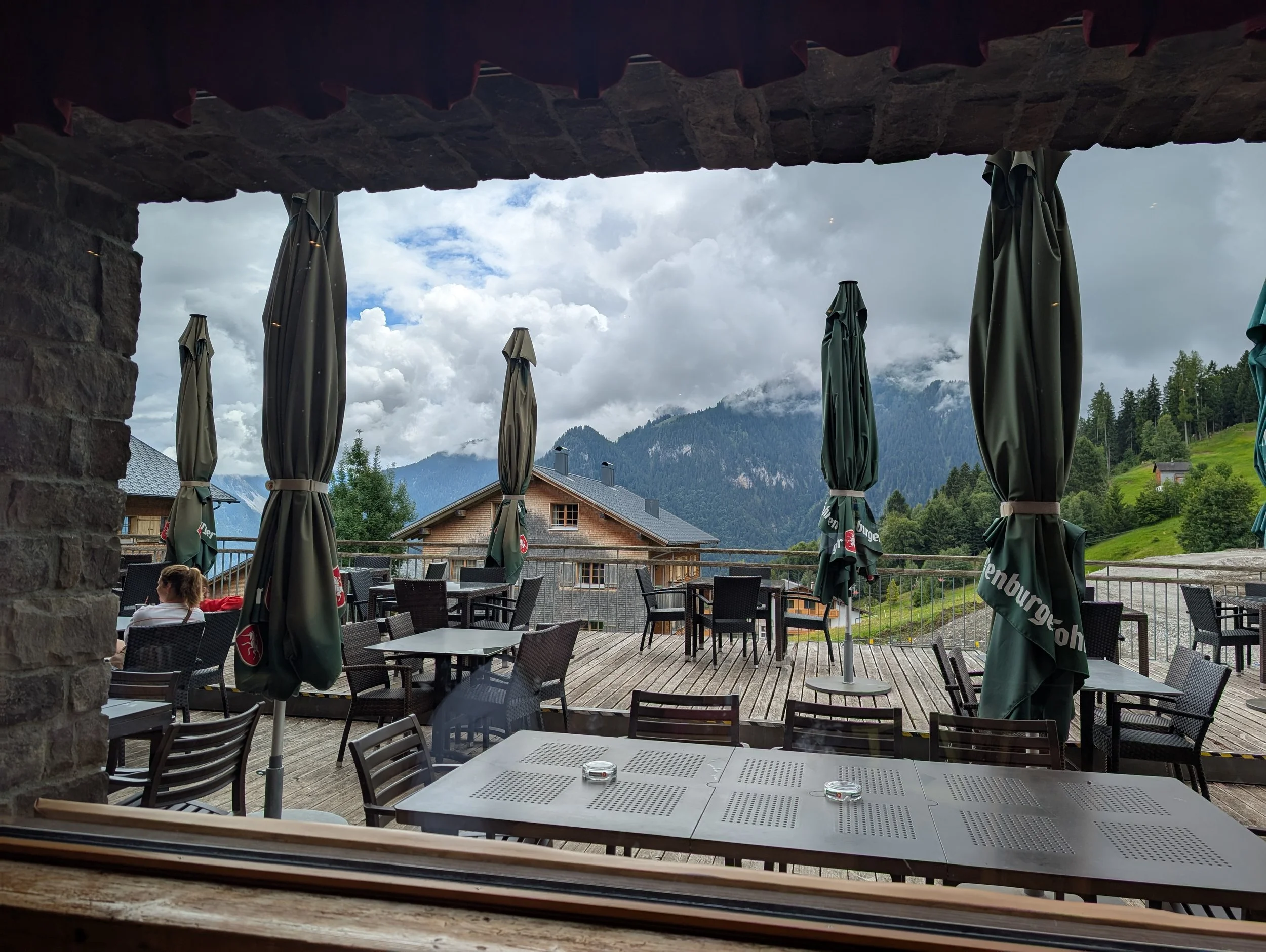 Blick von einem Innenraum auf eine Terrasse mit Tischen und Stühlen, umgeben von bewölktem Himmel, Bergen und Wald im Hintergrund.