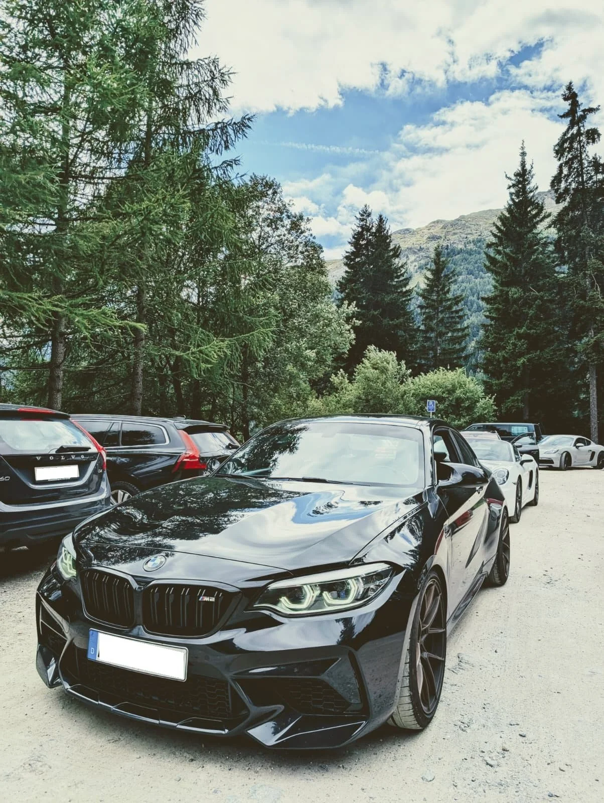 Schwarzer BMW M3 Autos in einem Parkplatz vor einer Waldkulisse mit Bäumen und Bergen unter einem blauen Himmel.