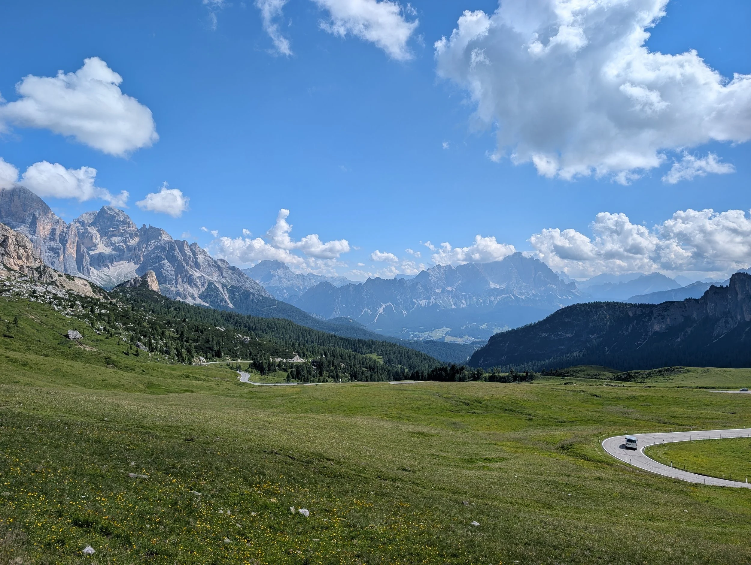 Berglandschaft mit grünen Wiesen, einer kurvigen Straße und Bergen im Hintergrund unter einem bewölkten Himmel.