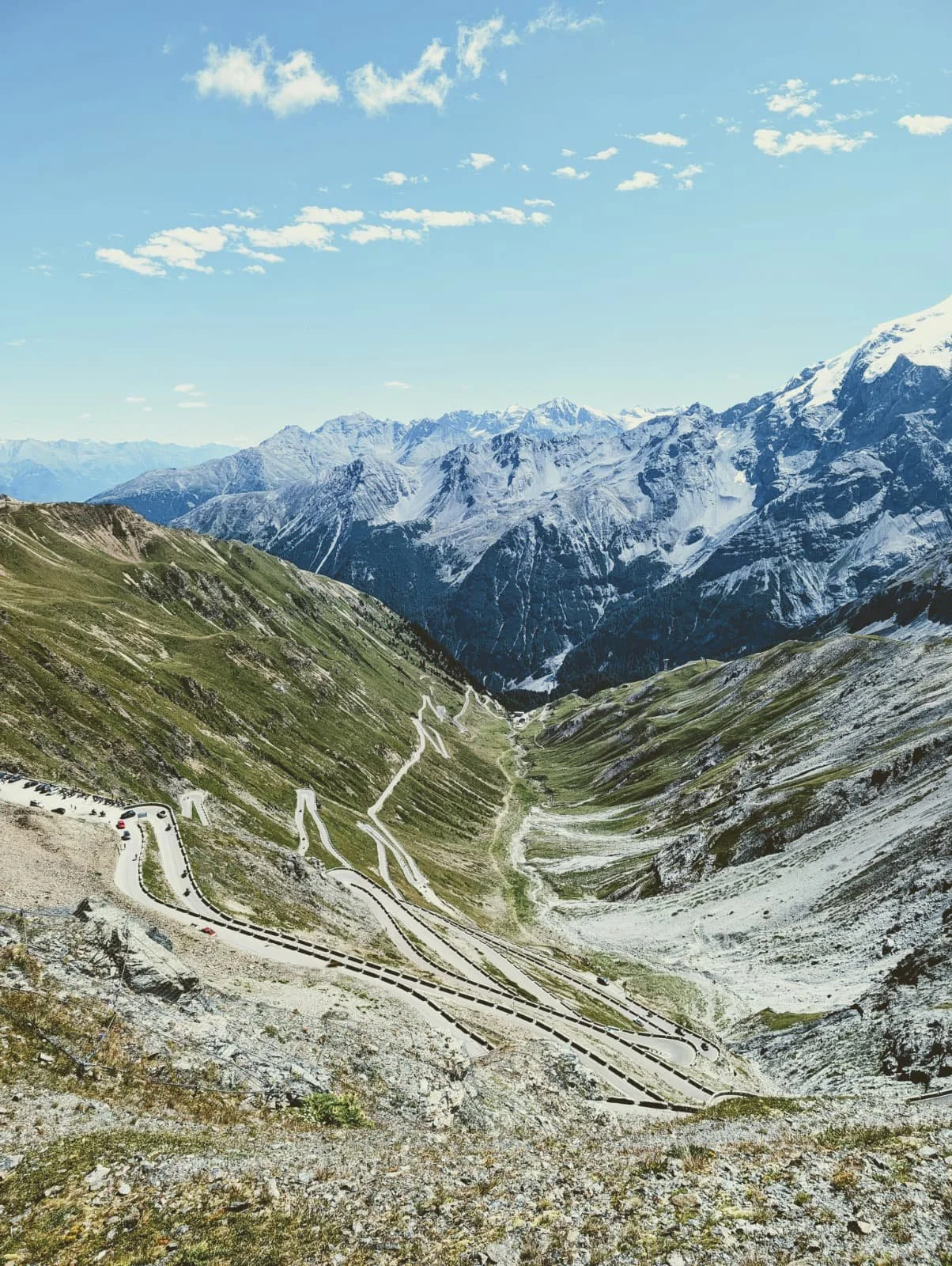 Berglandschaft mit schneebedeckten Gipfeln, grünen Hängen und kurvigen Straßen in den Alpen unter blauem Himmel mit Wolken