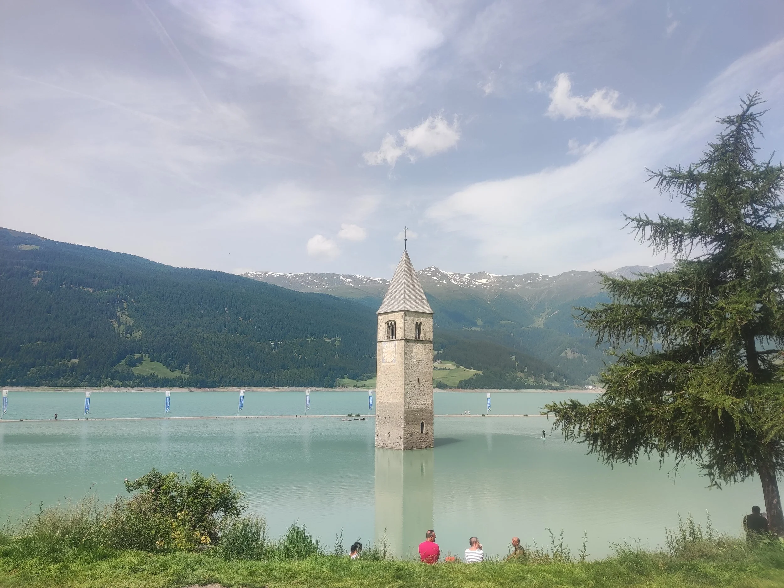 Der Kirchturm der Kirche inmitten eines Sees mit Bergen im Hintergrund, einige Bäume und Menschen am Ufer