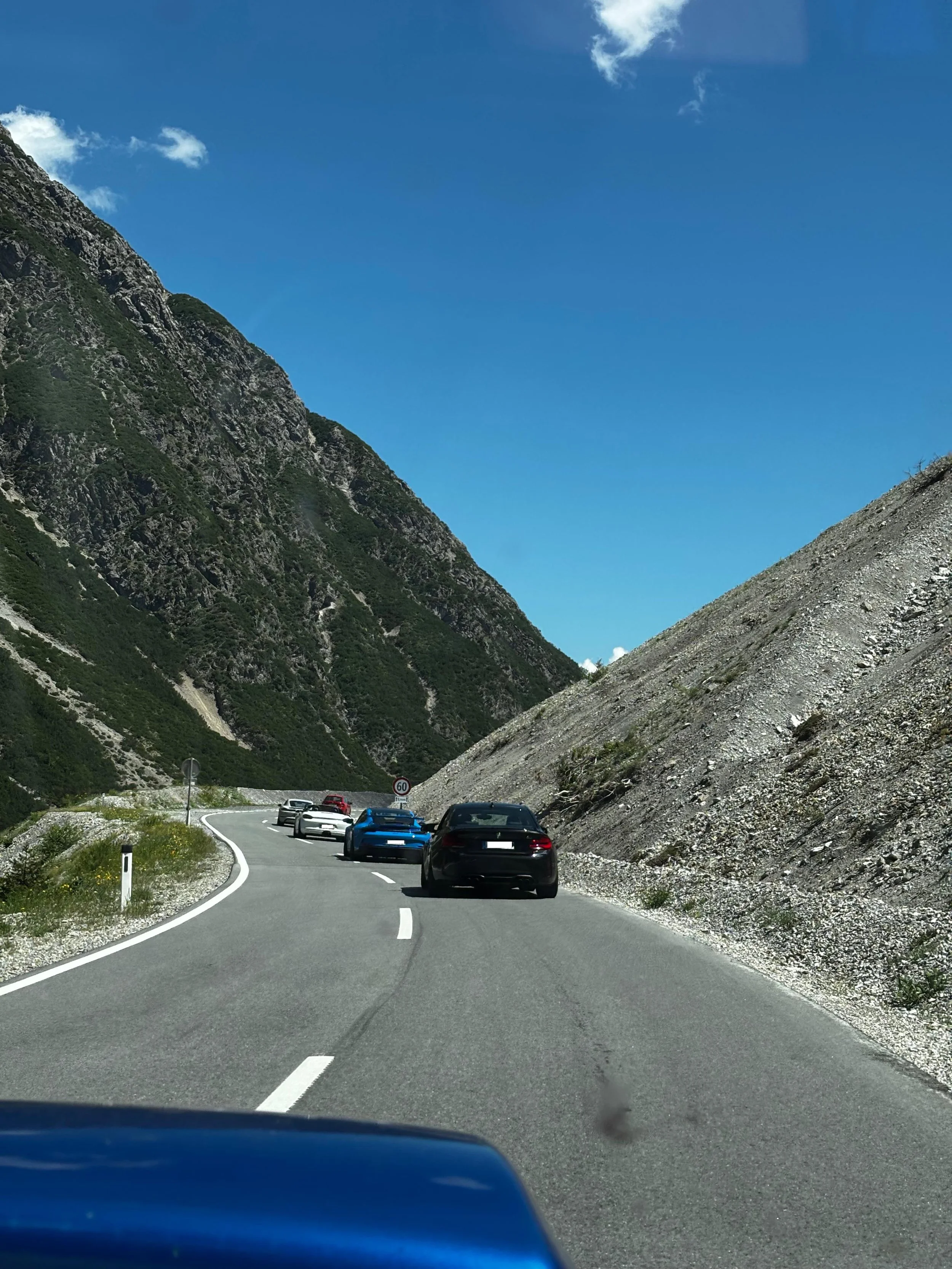 Autofahrt durch eine kurvige Bergstraße mit mehreren Autos unterwegs, umgeben von hohen Bergen bei blauem Himmel.