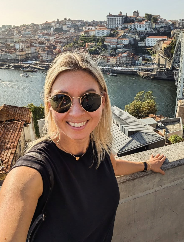 Eine lächelnde junge Frau mit Sonnenbrille macht ein Selfie auf einer Brücke in Porto, Portugal, mit Blick auf den Douro-Fluss und die Stadt mit Hügeln und historischen Gebäuden im Hintergrund.