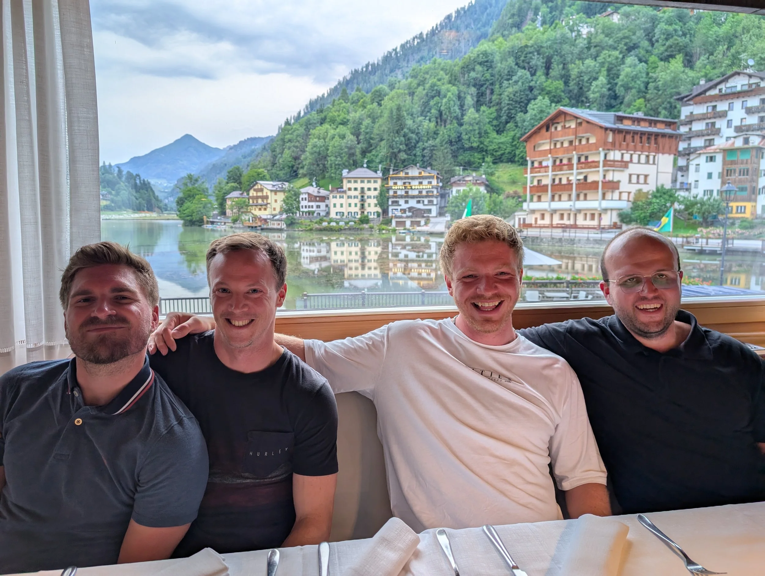 Vier Männer sitzen an einem Tisch im Restaurant, mit Blick auf einen Fluss, bunte Häuser und grüne Hügel im Hintergrund, bei schönem Wetter.