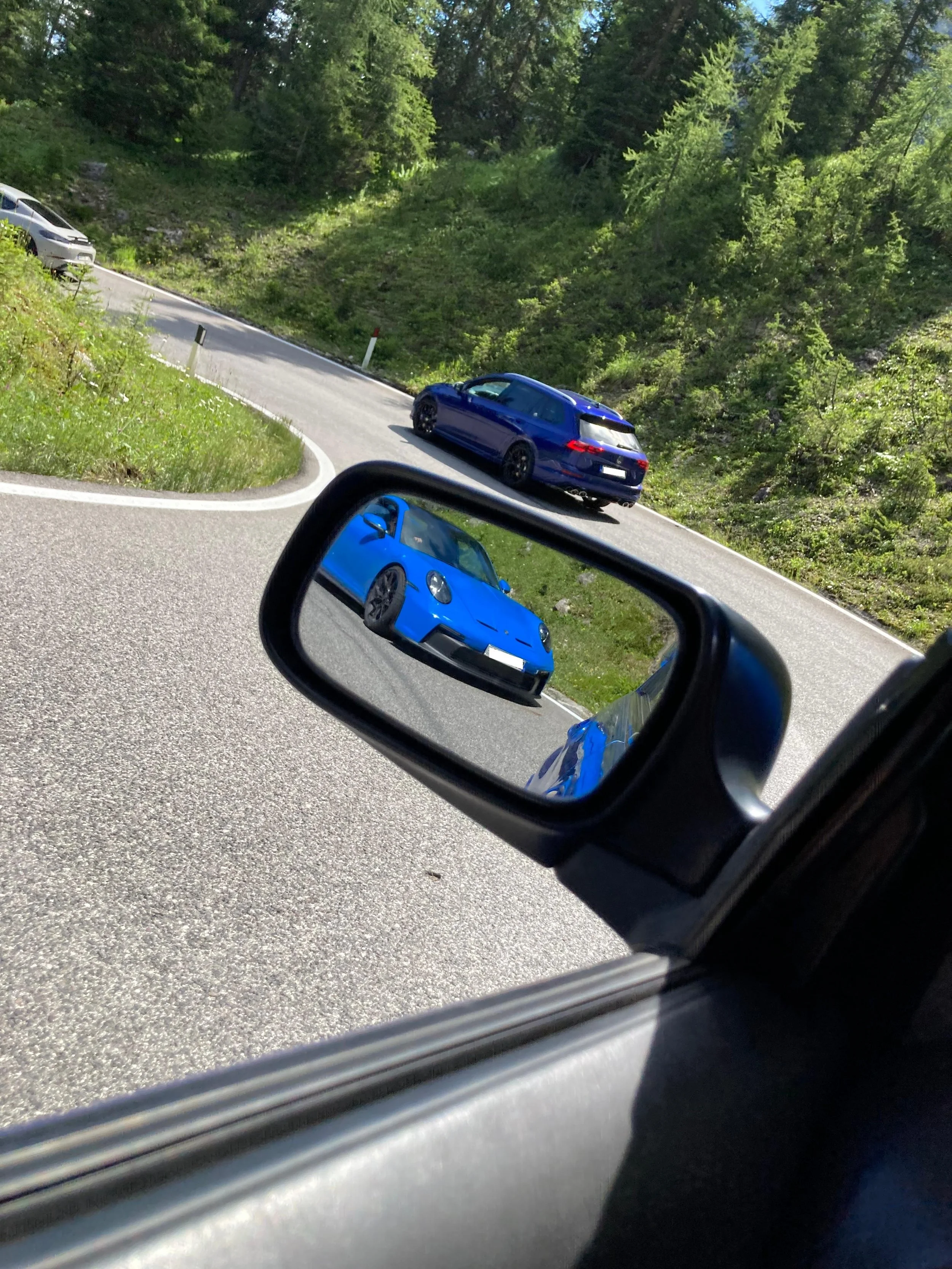 Blick in den Rückspiegel eines Autos auf eine kurvige Straße in einer grünen bergigen Landschaft mit drei blauen Autos, die die Straße hochfahren.