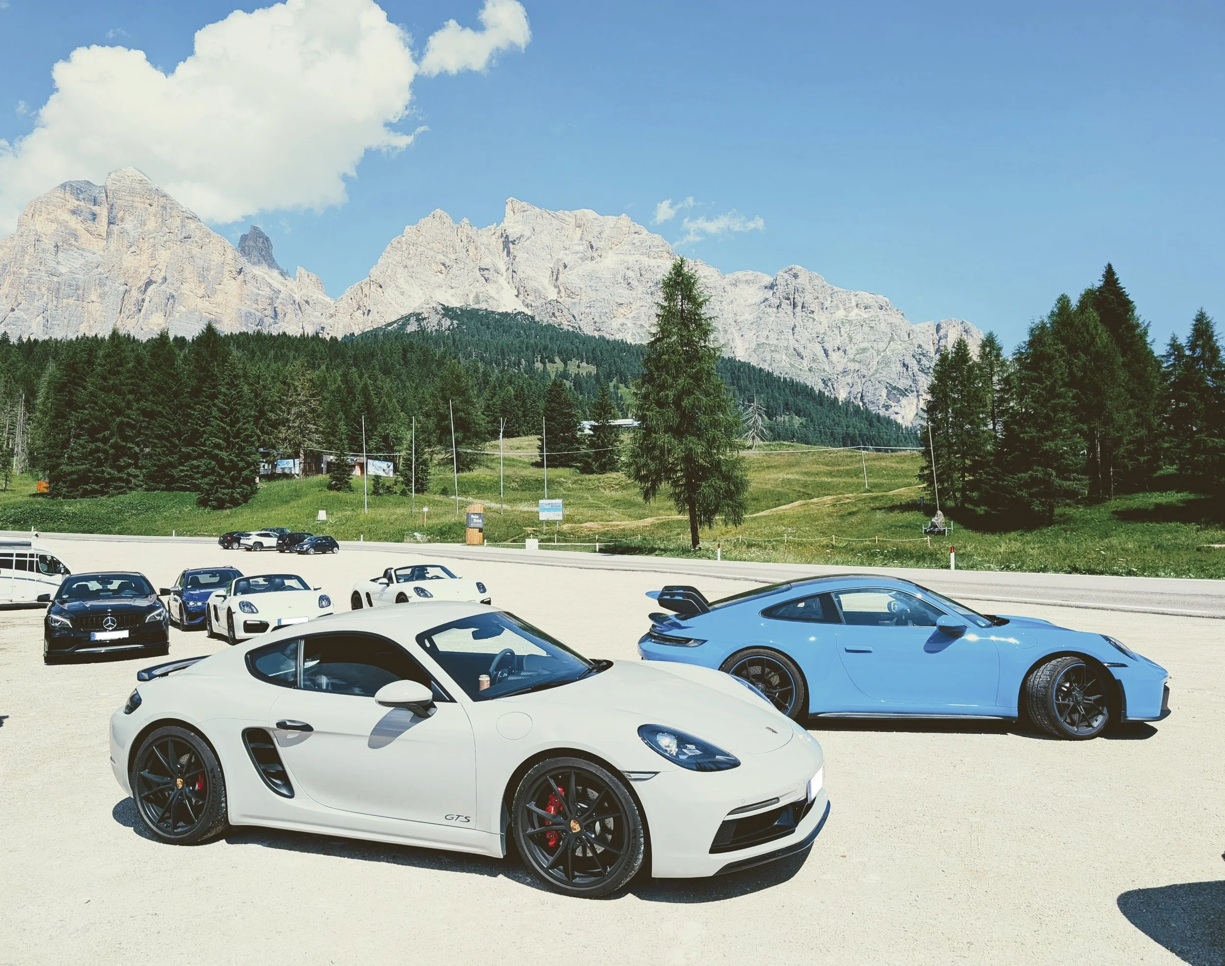 Mehrere Sportwagen, darunter ein weißer Porsche Cayman und ein blauer Porsche 718, stehen in einem Parkplatz vor einer Berglandschaft mit Bäumen und Bergen im Hintergrund bei sonnigem Wetter.