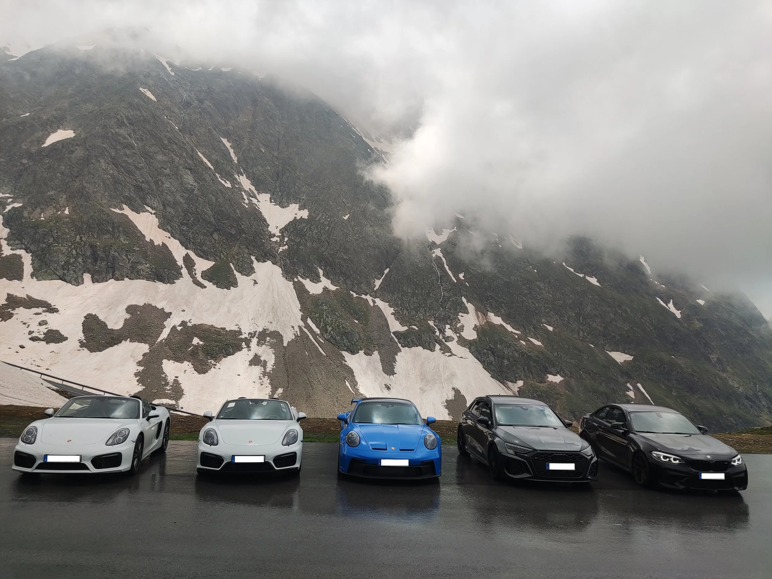 Fünf Sportwagen, darunter zwei Porsche, auf nasser Straße vor einer bergigen, bewölkten Landschaft.