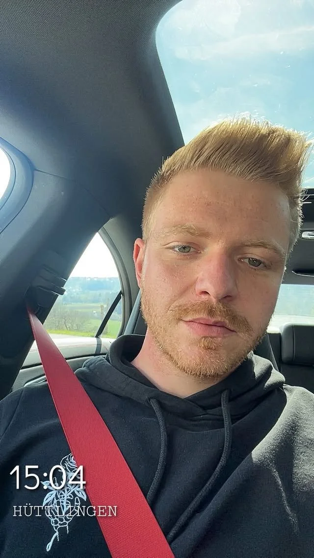 Ein junger Mann mit blondem Haar und leichtem Bart sitzt im Auto, trägt ein schwarzes Hoodie mit einem weißen Design auf der Brust, hat ein rotes Sicherheitsgurt an und schaut in die Kamera.