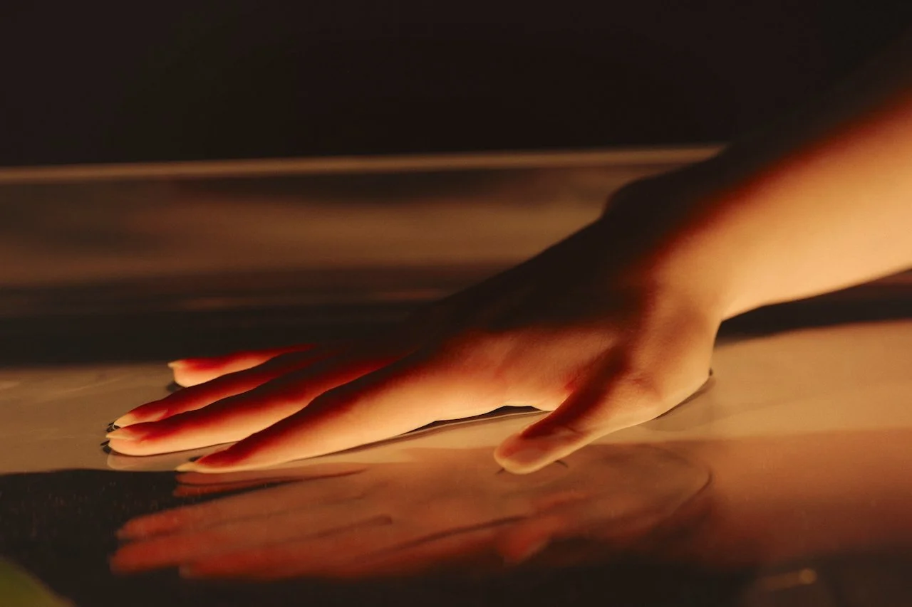A person's hand is touching a reflective, possibly metallic or glass surface, with soft, warm lighting creating a gentle glow on the skin.