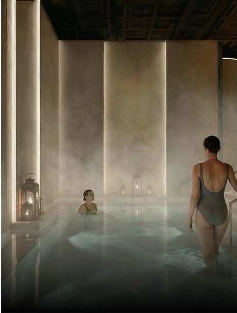 Two women in swimsuits relax in an indoor hot spring or bath with stone walls and lanterns, creating a serene atmosphere.