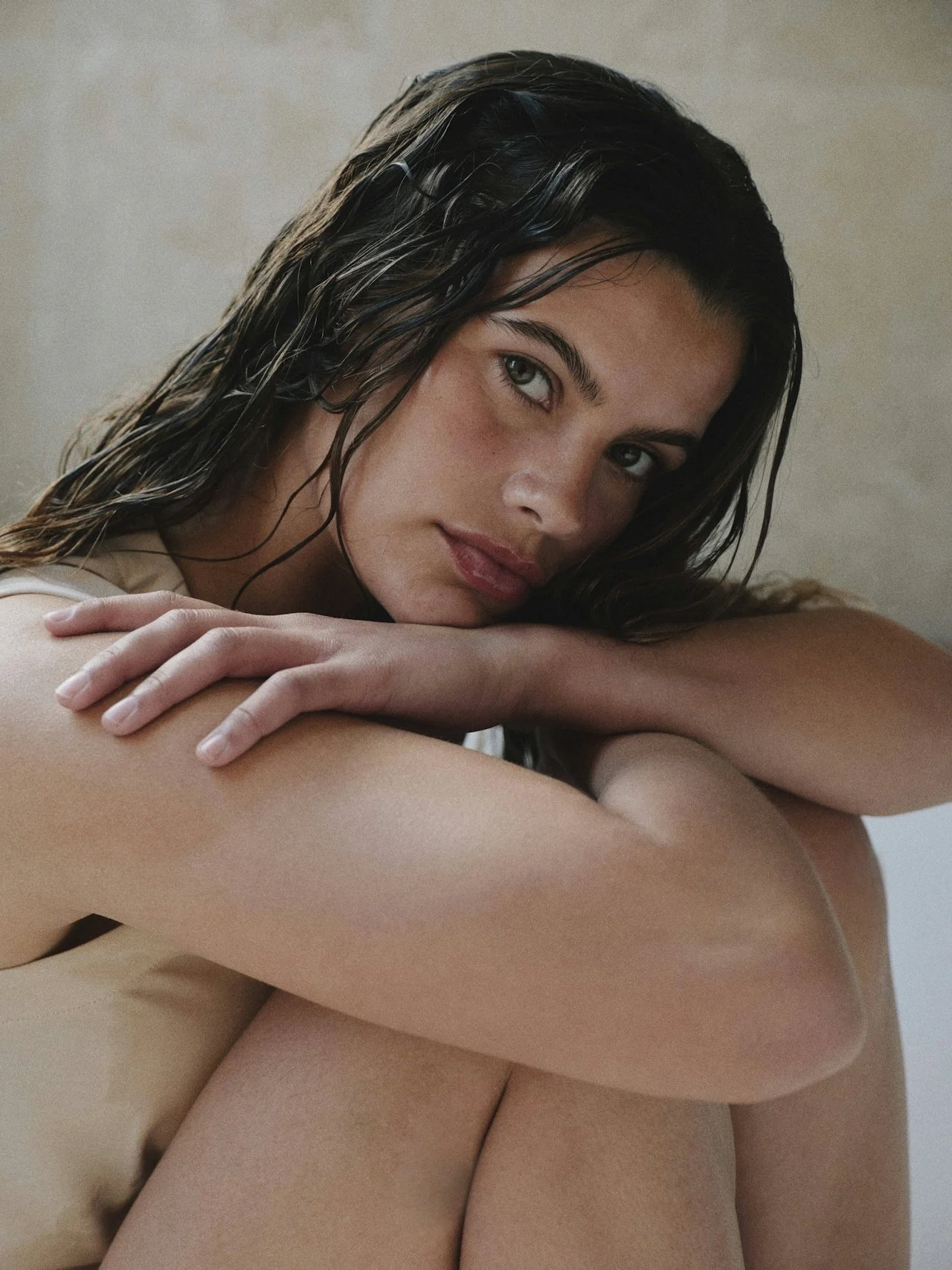 Close-up of a woman with wet dark hair and blue eyes resting her head on her knees, looking into the camera.