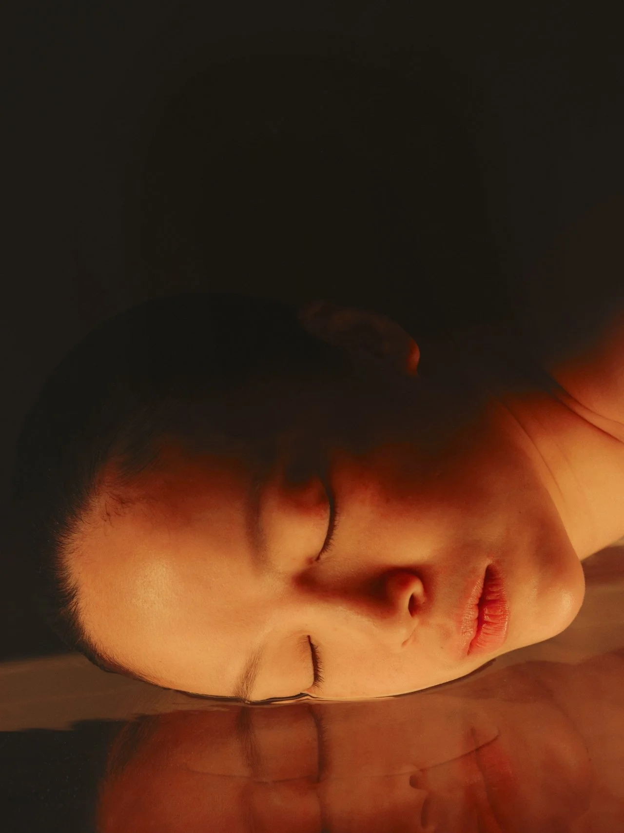Close-up of a woman with her eyes closed, lying on a reflective surface with a dark background.