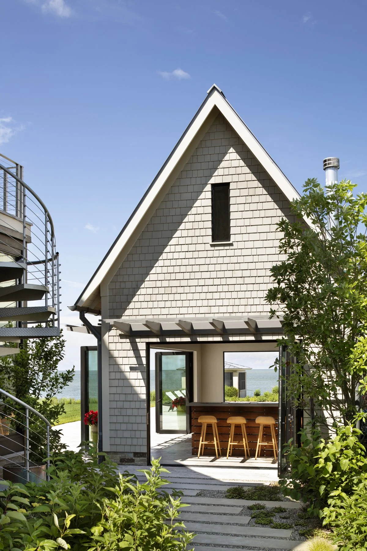 Modern house with shingle exterior, large windows, and an outdoor bar area with three stools, surrounded by greenery and overlooking the ocean under a blue sky.