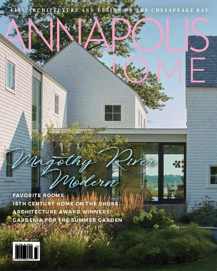 Kimmel Studio Architects Annapolis Home magazine with a modern white house with blue siding, large windows, surrounded by greenery and trees, and a view of the water in the background.