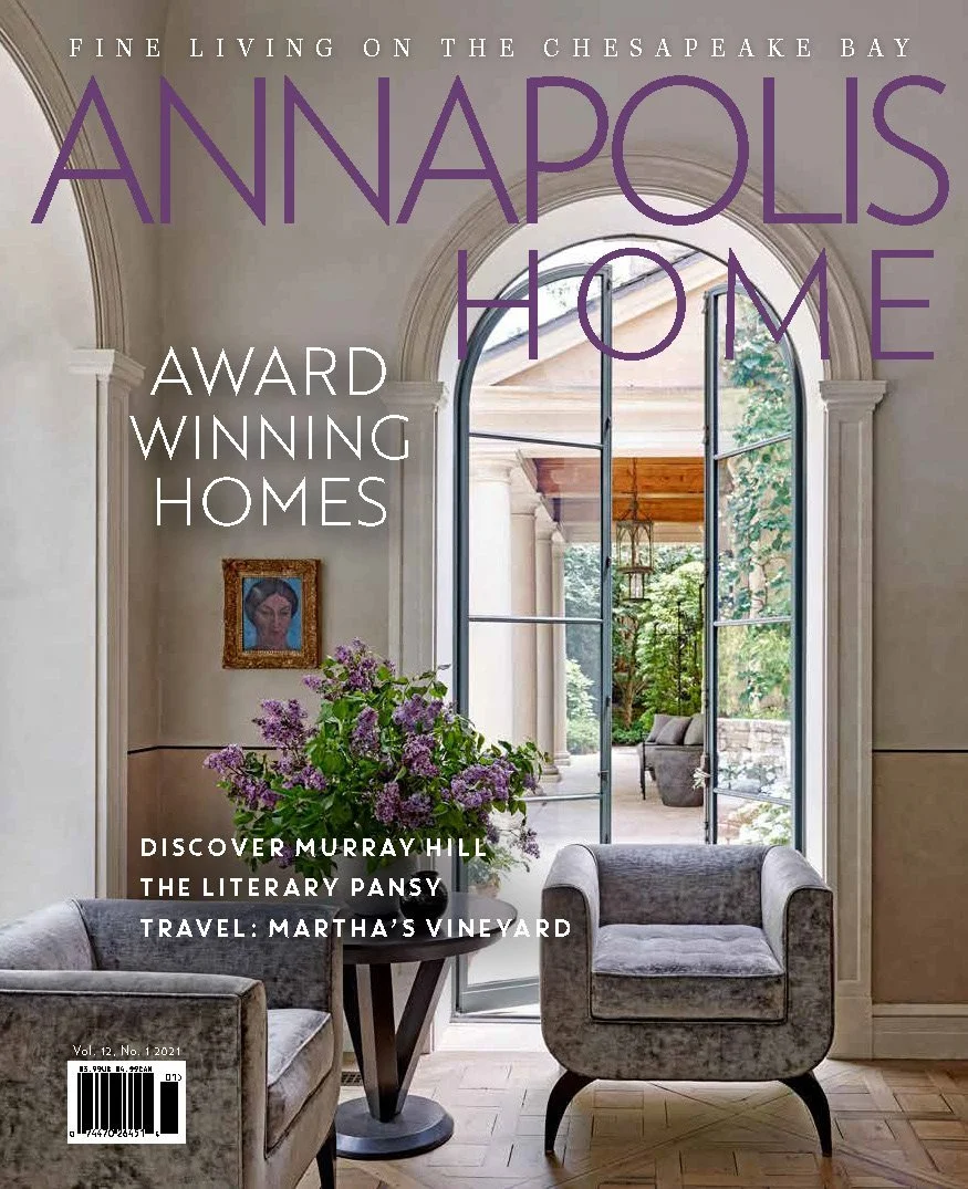 Kimmel Studio Architects Annapolis Home Magazine, featuring a bright, elegant interior room with large arched window, light-colored walls, purple flowers on a table, and cozy furniture, with a view of an outdoor patio covered porch.
