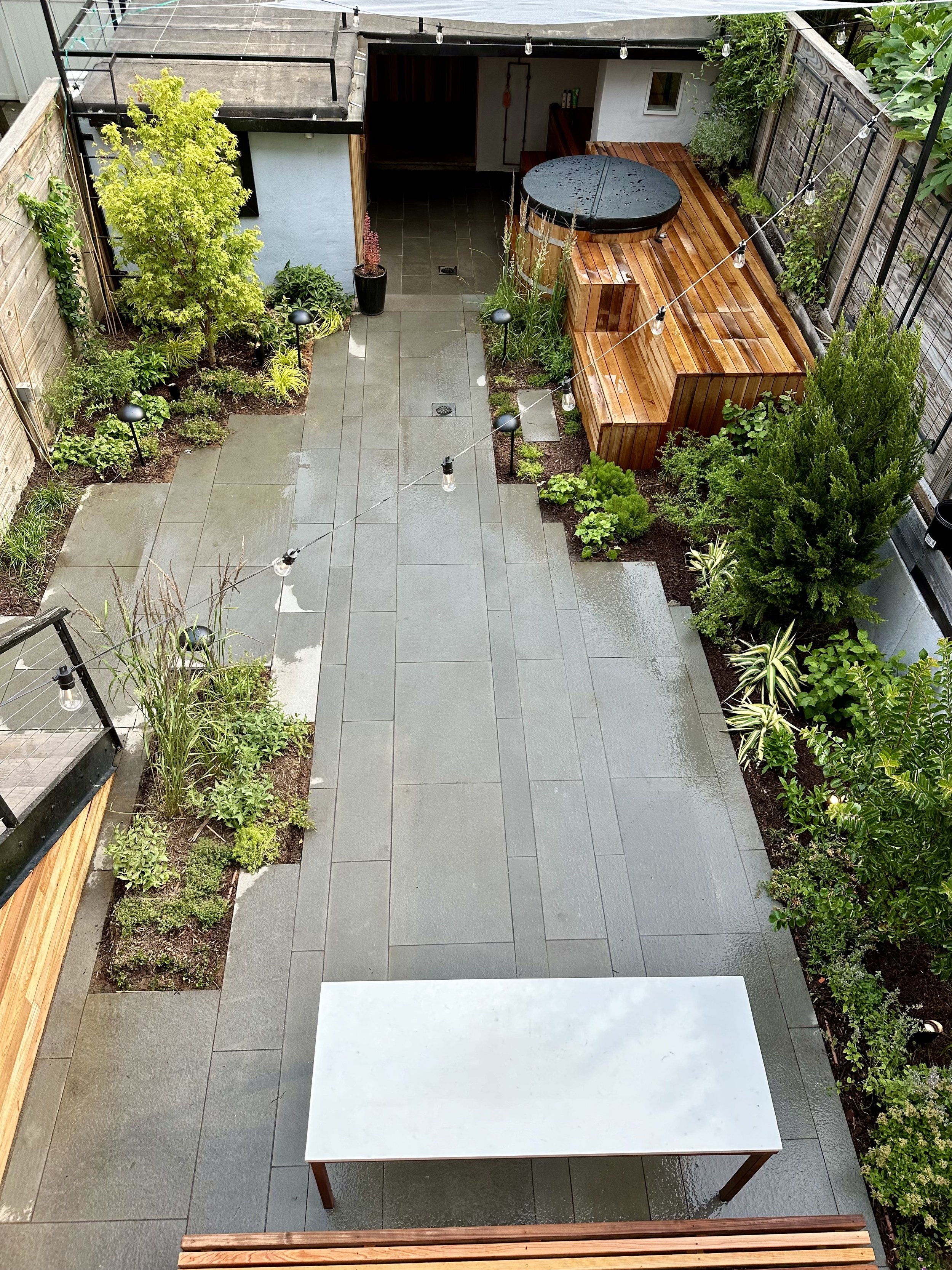 Rain-soaked backyard patio with gray tile flooring, a large white table, string lights overhead, a wooden hot tub, and various green plants and trees along the perimeter.
