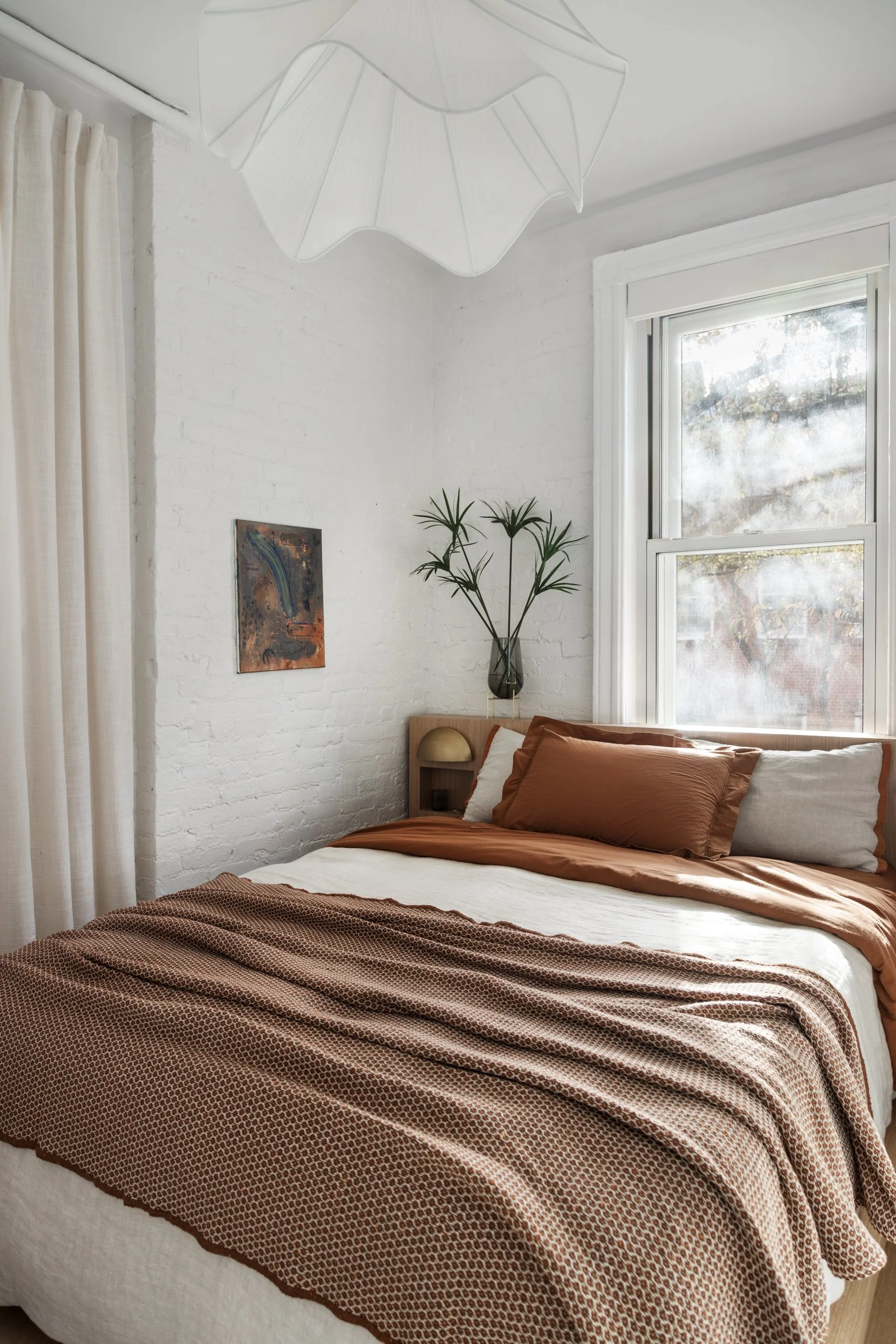 A neatly made bed with rust-colored pillows and a patterned brown blanket in a bright bedroom with white brick walls, a window with a view, a small nightstand with a lamp, a tall plant, and a hanging white pendant light.
