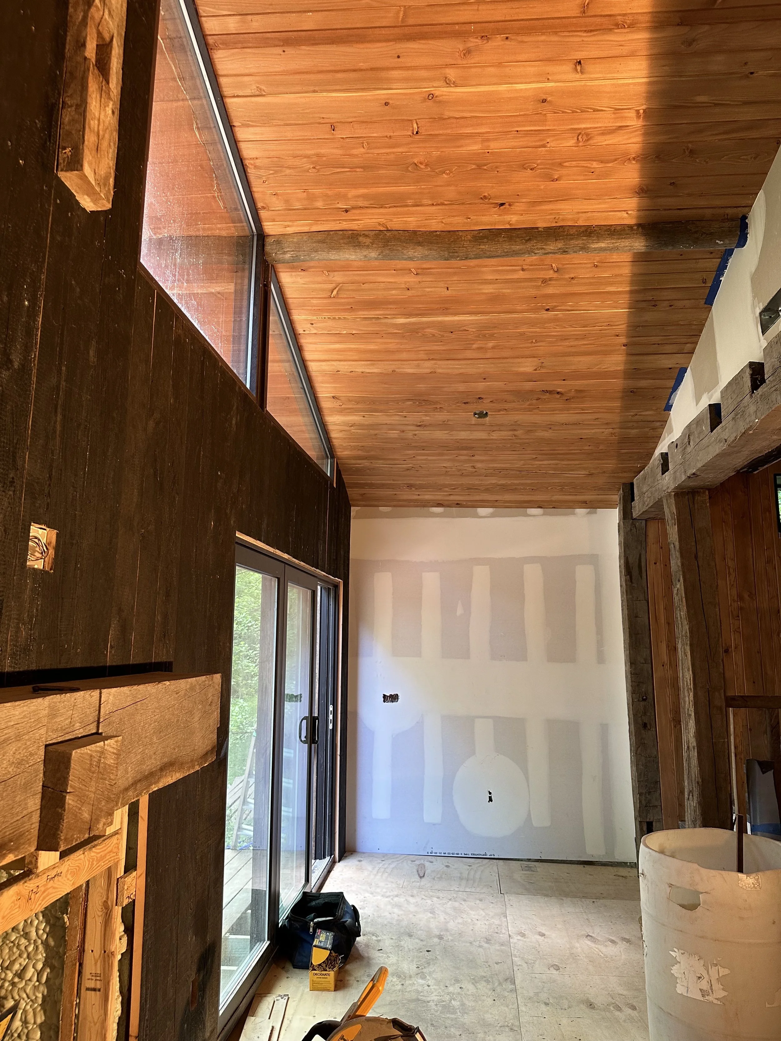 Interior room under construction with unfinished drywall, wood-paneled ceiling, sliding glass door, and construction tools and materials on the floor.