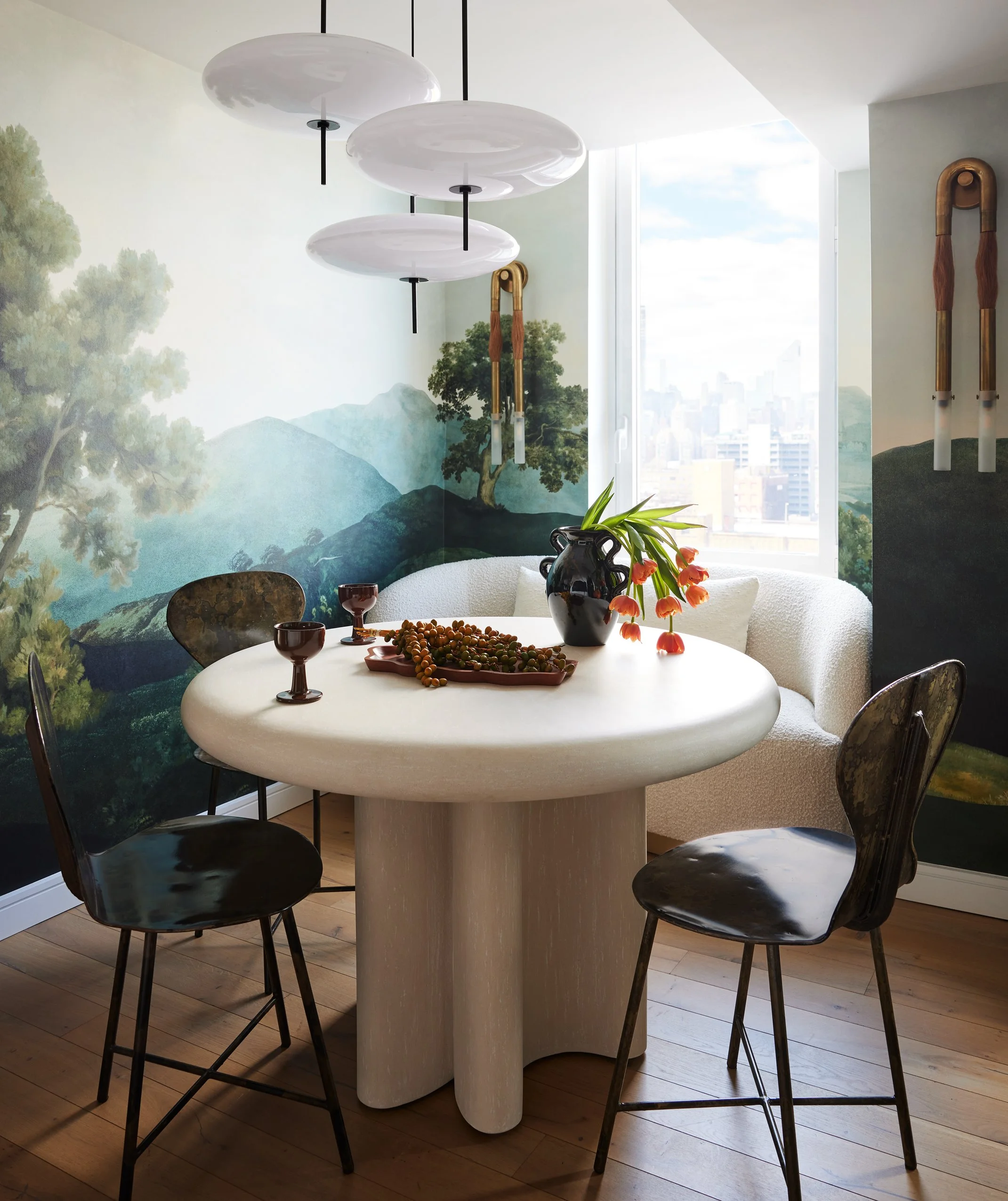 Dining room with a round, white table, three black chairs, a white curved sofa, a mural of mountains and trees on the wall, a black vase with flowers, grapes, and two cups on the table, and artistic lighting fixtures hanging from the ceiling.