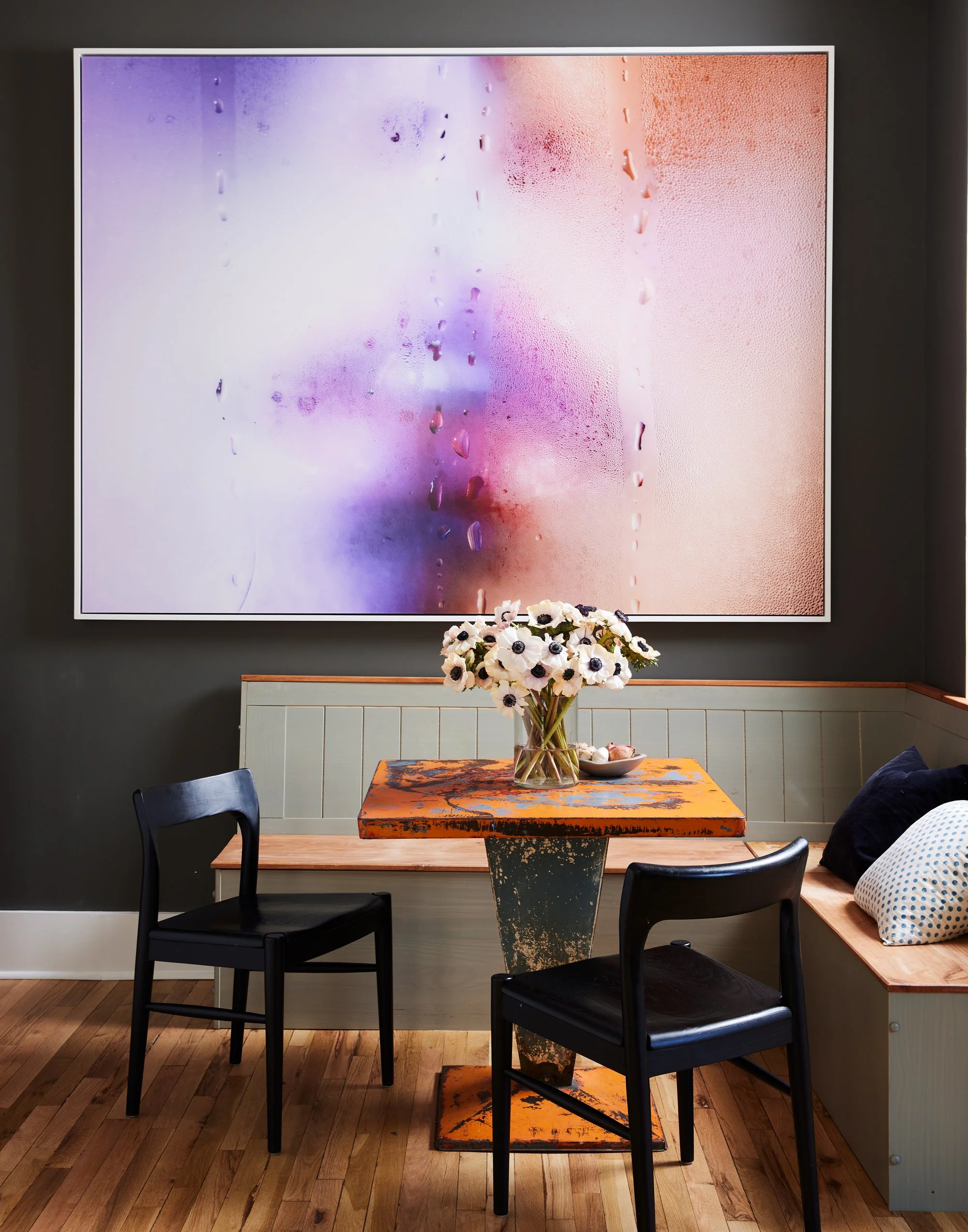A cozy dining area with a round wooden table featuring a distressed orange and black surface, decorated with a glass vase holding white flowers with black centers. Two black chairs are placed around the table, and there's a built-in bench with pillows against a dark wall. A large, colorful abstract artwork with water droplets on its surface hangs behind the table.