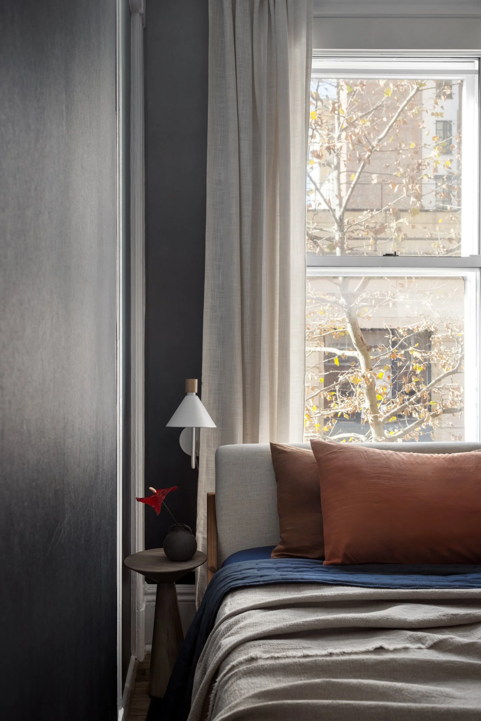 A cozy bedroom corner with a bed, brown pillows, a nightstand with a small vase and flower, a wall lamp, and a window with beige curtains letting in natural light.