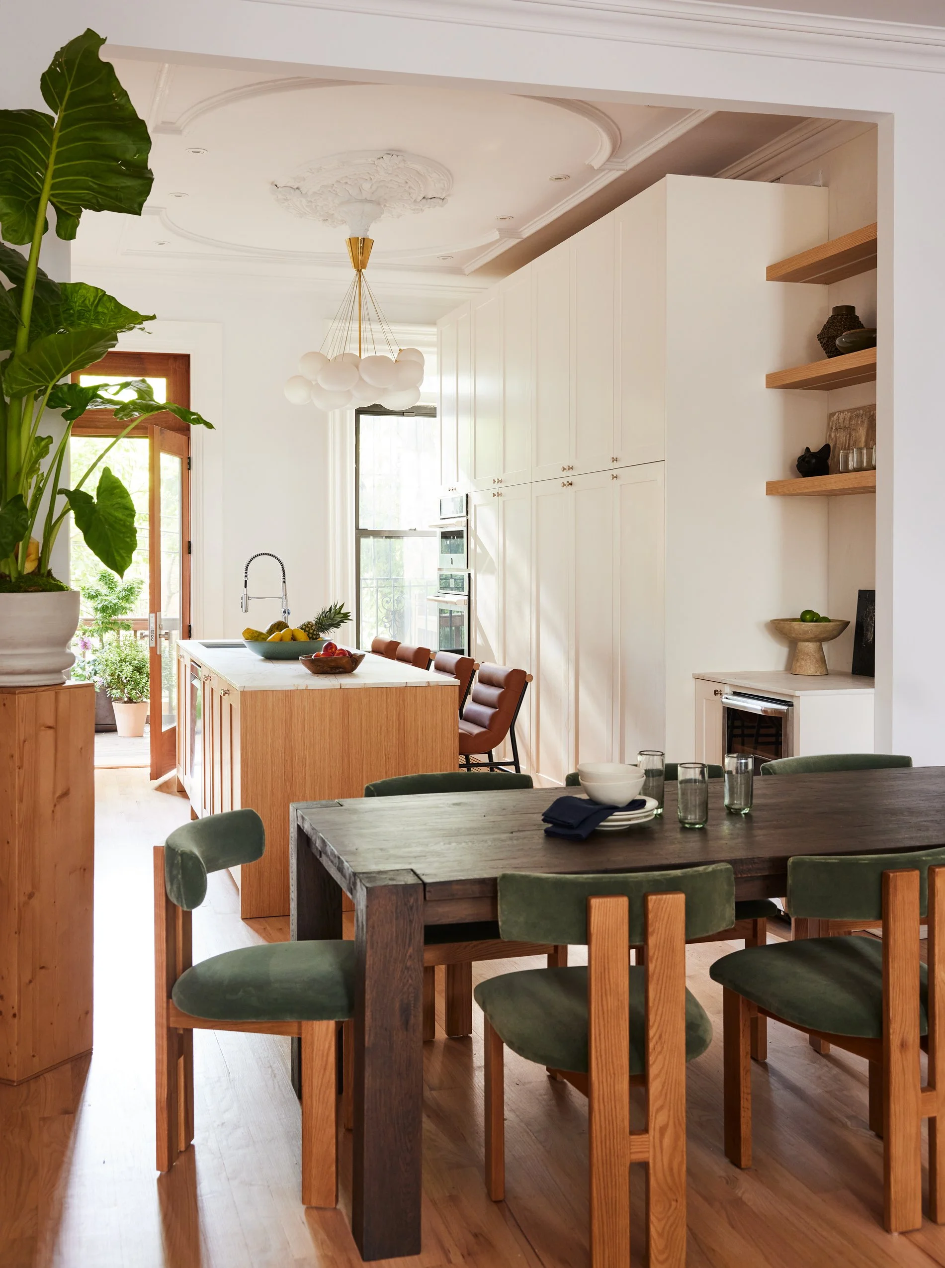 A dining area with a dark wooden table and six green velvet chairs. Behind it, a kitchen island with a white countertop and bar stools. The kitchen has white cabinets, a window, and open wooden shelves with decor. A large leafy plant is to the left.