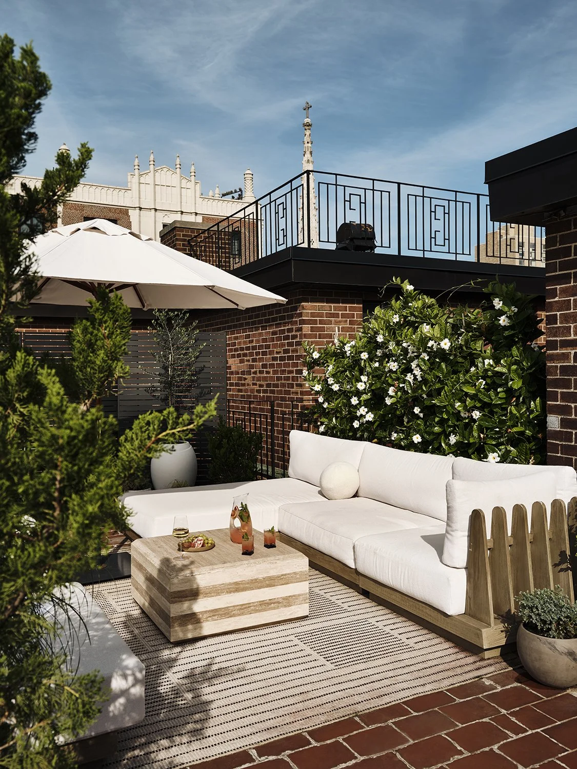 Outdoor rooftop patio with a white cushioned sectional sofa, a wooden coffee table with drinks and snacks, potted plants, a large white umbrella, brick walls, and a decorative iron railing, with a blue sky above.