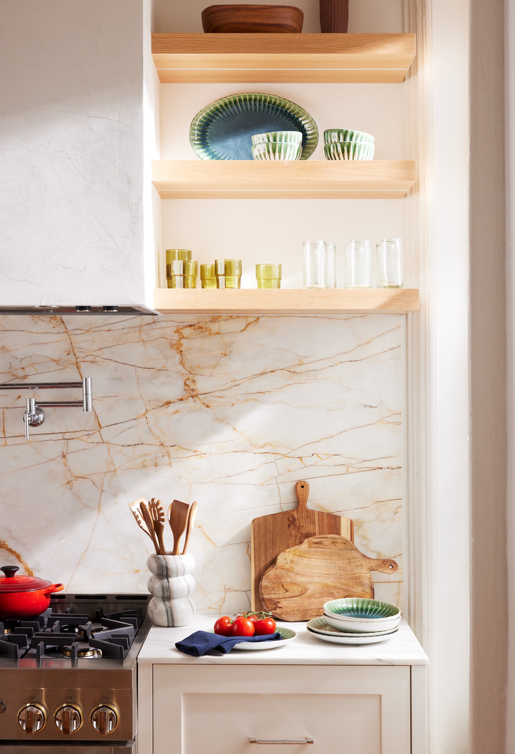 A kitchen with a marble backsplash, wooden cutting boards, a pot with tomatoes, a blue napkin, and dishes on a white countertop. Open wooden shelves above hold green and clear glasses and dishes.