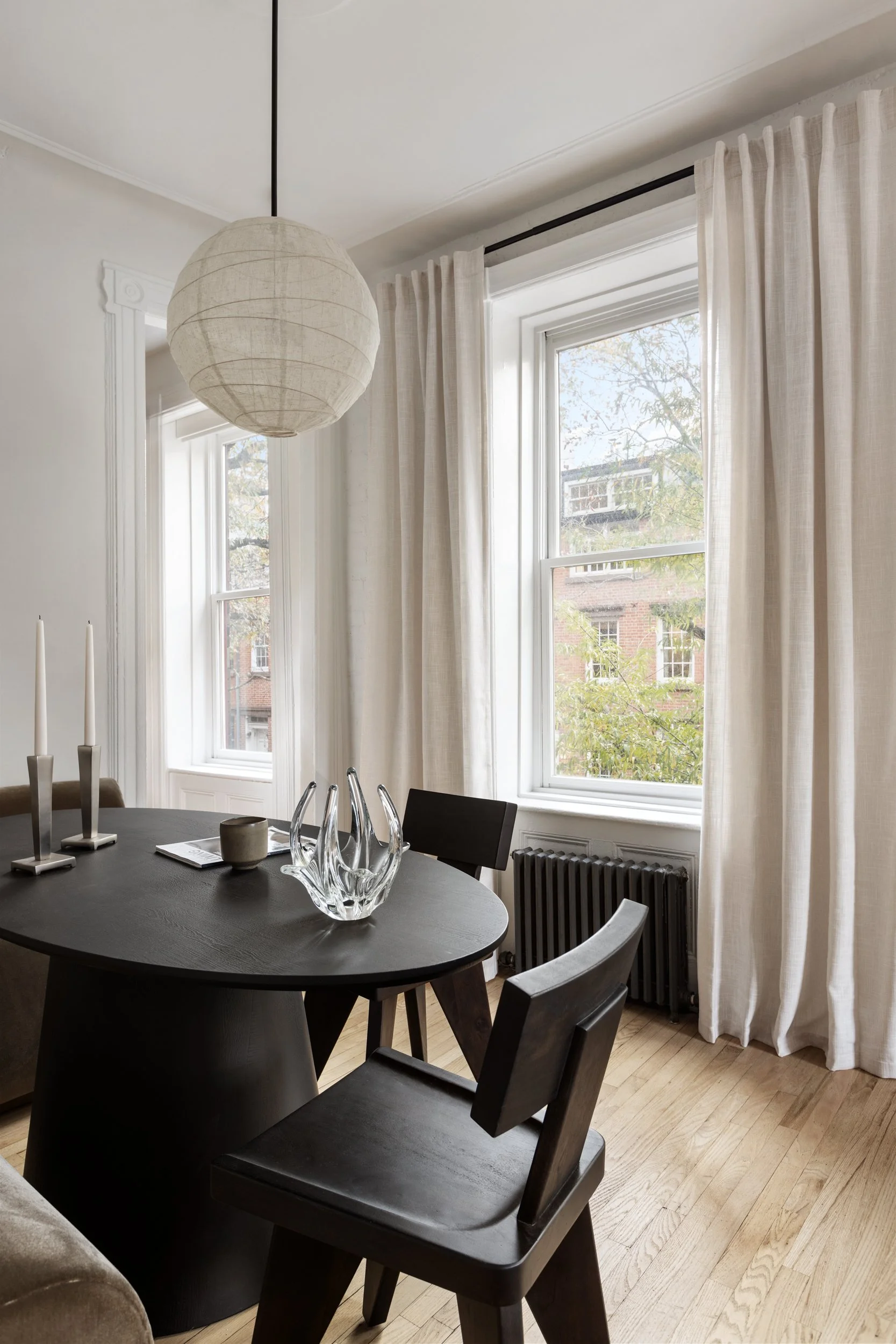 A dining room with large windows and white curtains, a black oval dining table with decorative glassware and candles, surrounded by black chairs, and a paper lantern hanging from the ceiling.