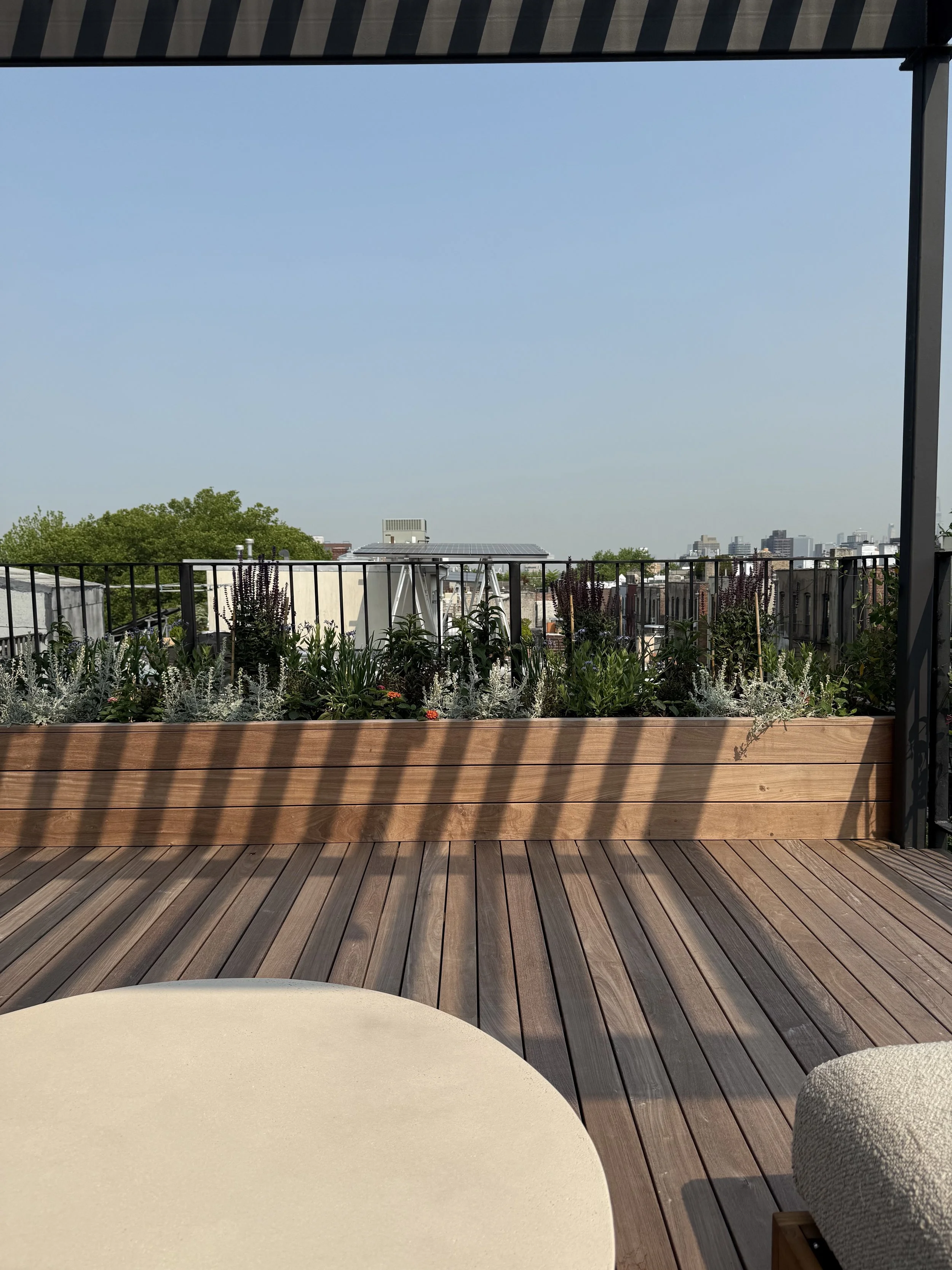 View of a rooftop balcony with wooden flooring, a planter box with green plants and purple flowers, and city skyline in the background under a clear blue sky.