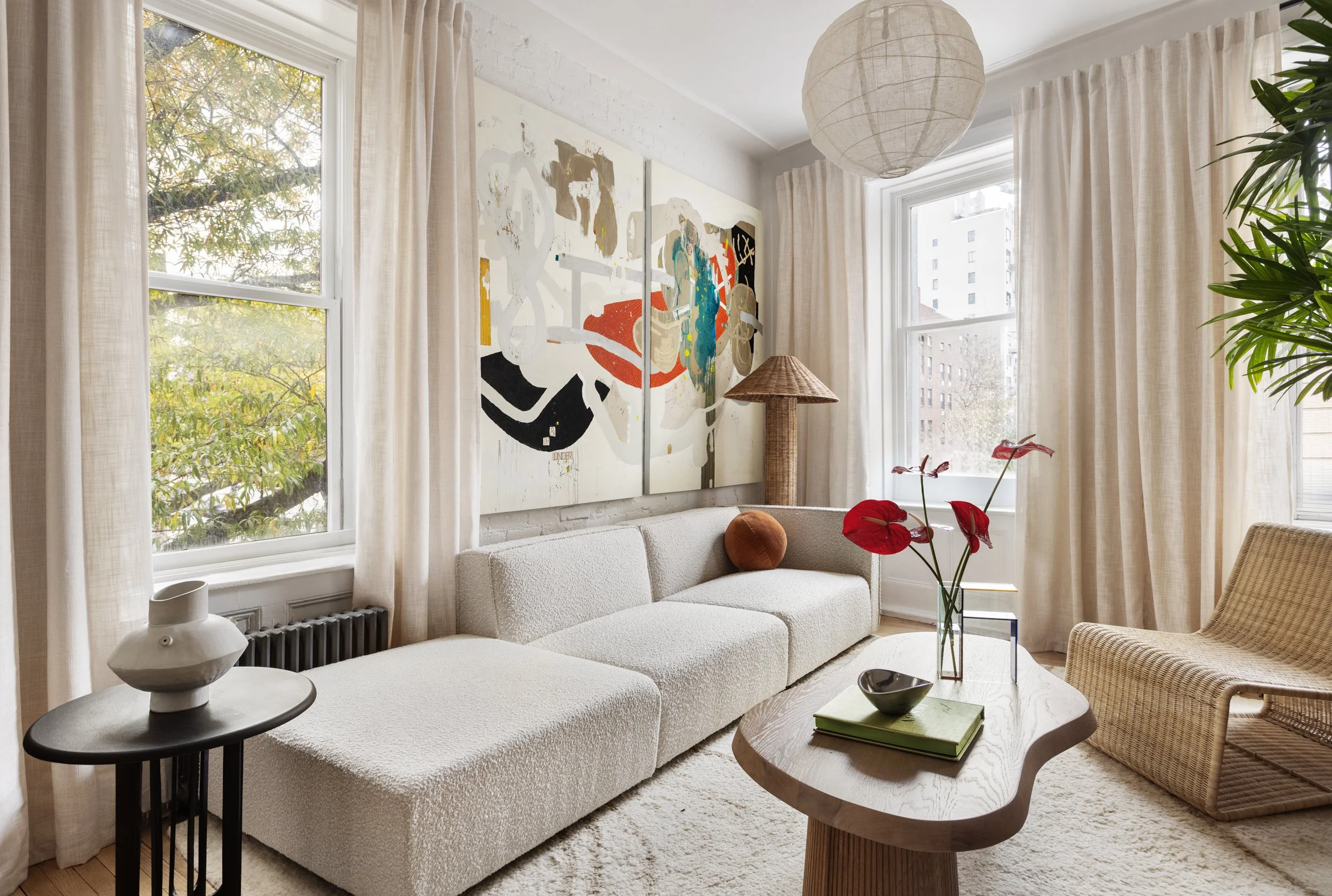 Bright living room with large windows, cream curtains, a white textured sofa, a wooden coffee table with a vase of red flowers, wall art, and a wicker chair.
