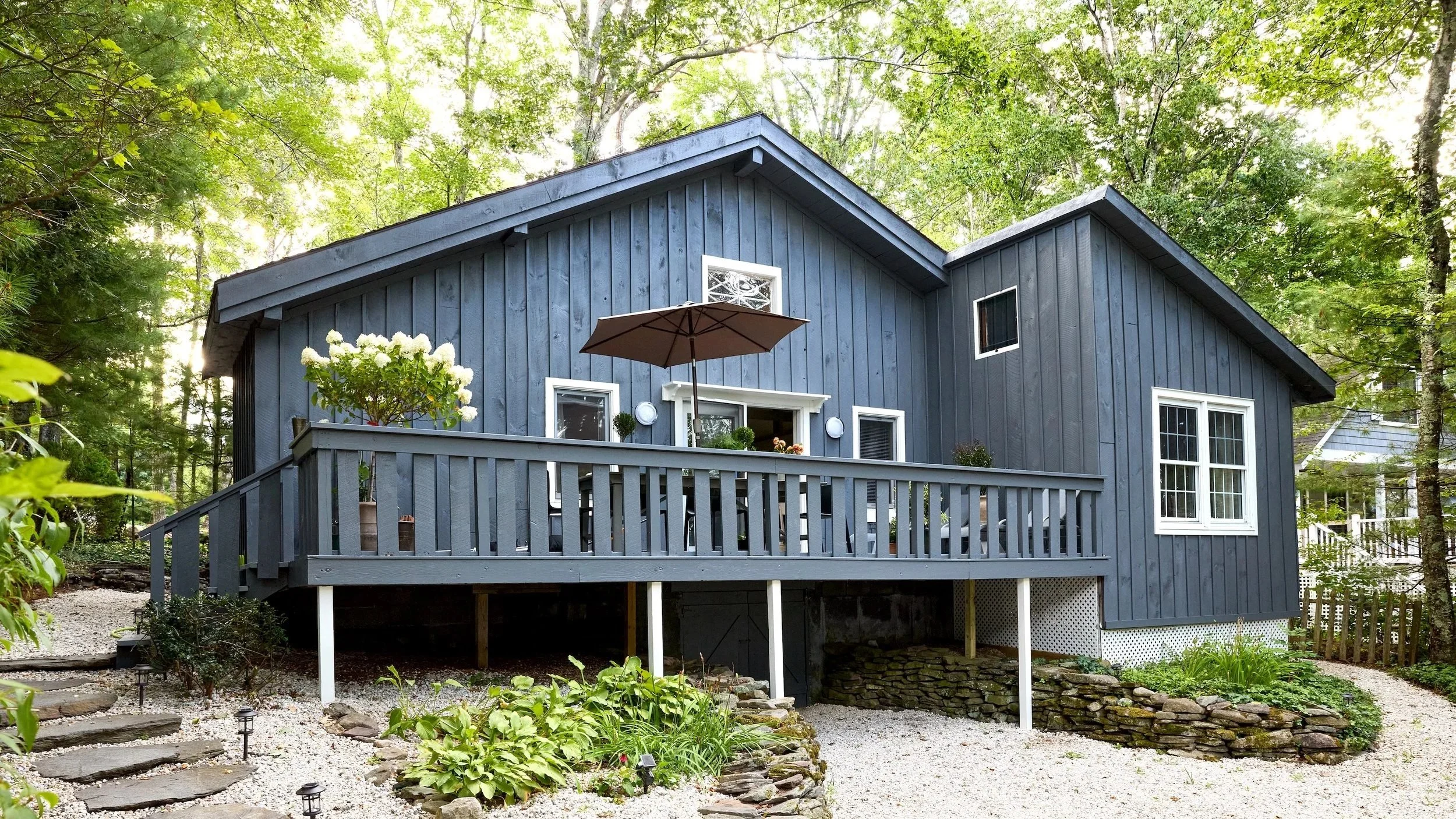 A blue wooden house on a hillside with a wooden deck, garden plants, potted flowers, a patio umbrella, surrounded by trees.