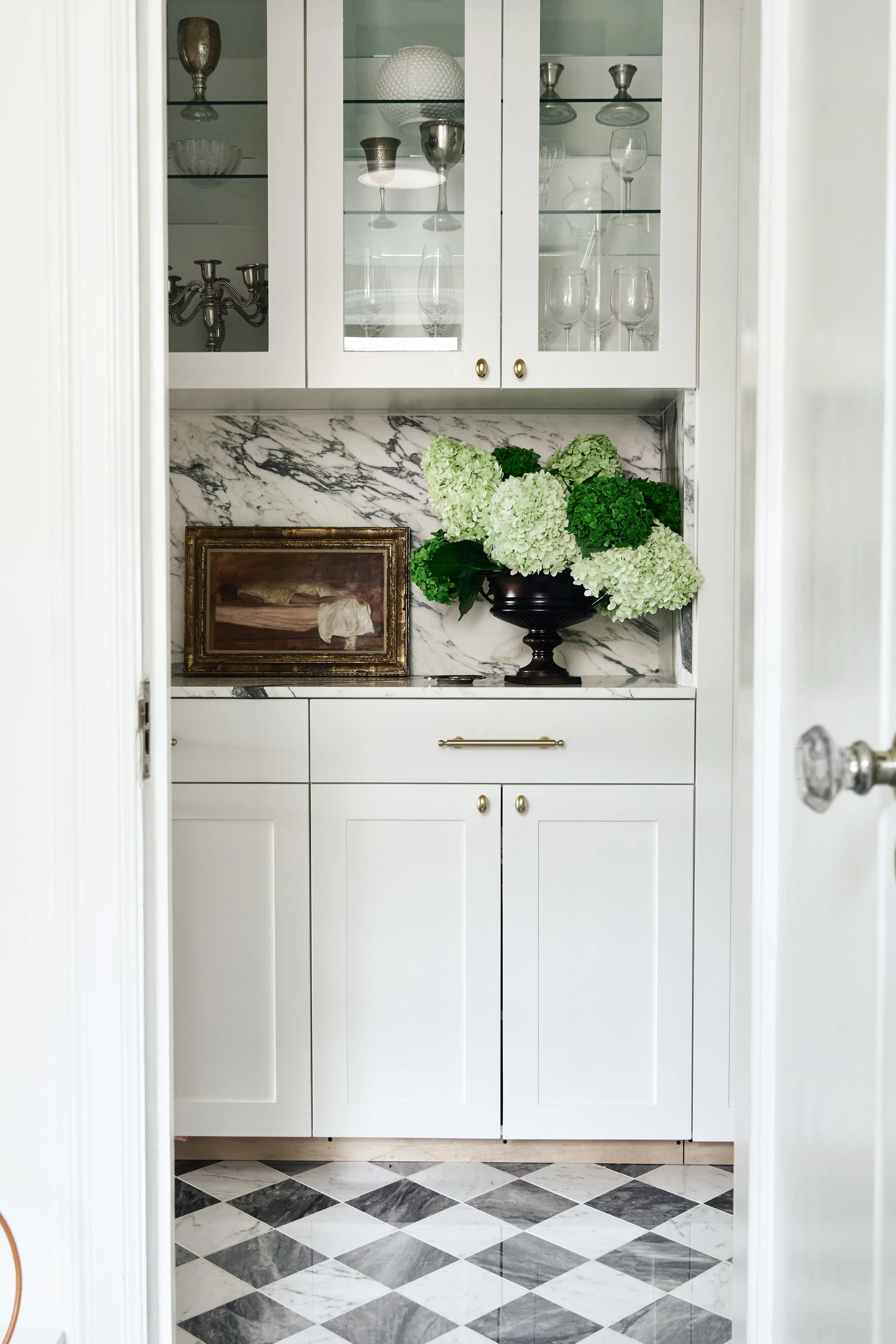 A white built-in cabinet with glass doors, marble backsplash, and patterned black and white checkered floor. Inside the cabinet are glassware and silver decorative items. On the counter, a large black vase with white and green hydrangeas, and a framed painting of a reclining woman.