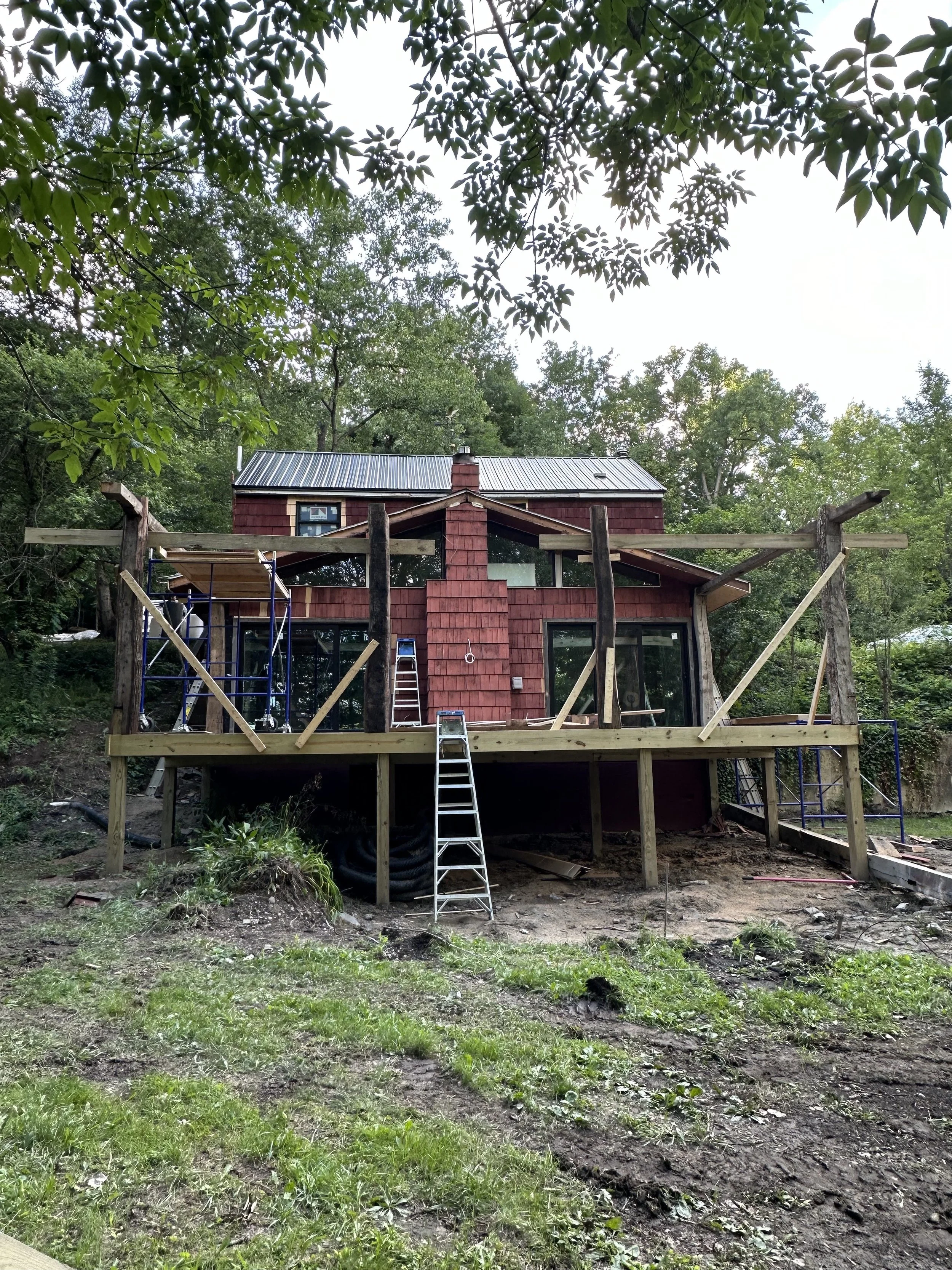 House under construction with a wooden deck, ladders, scaffolding, and surrounded by trees.